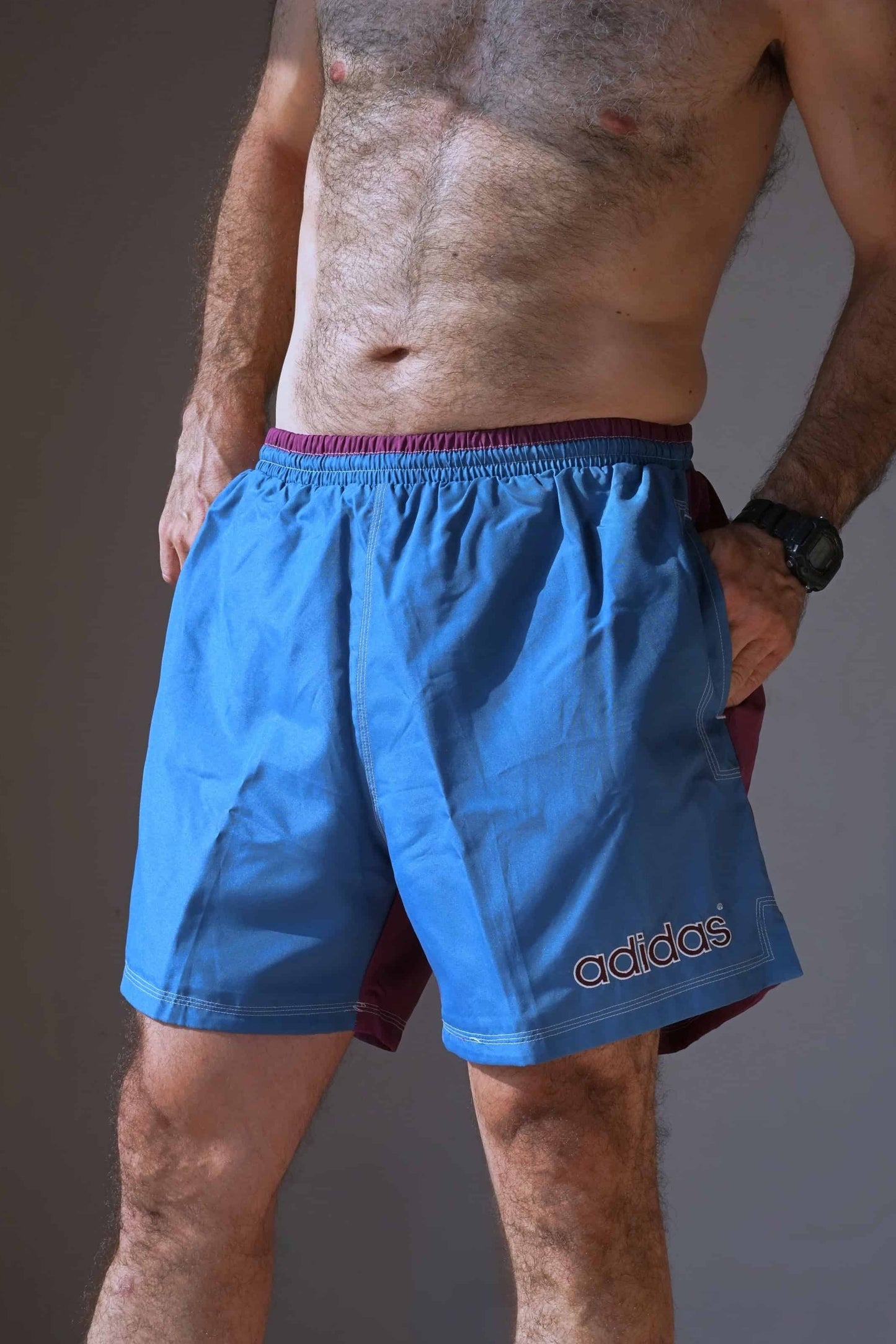 Man wearing vintage ADIDAS swim shorts with a blue front, purple back, and white stitched logo, standing with hands in pockets.