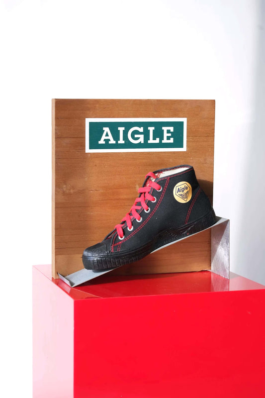 Angled view of vintage Aigle boot display. Wood back with logo, metal ramp, black canvas shoe with red laces on red base.