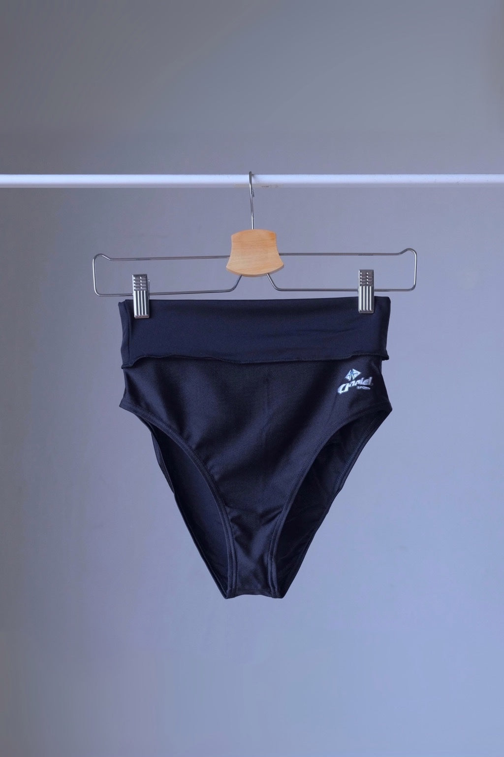 A pair of black, high-rise dance briefs with a folded-over waistband and a logo on the left hip, hanging on a metal hanger against a gray background.