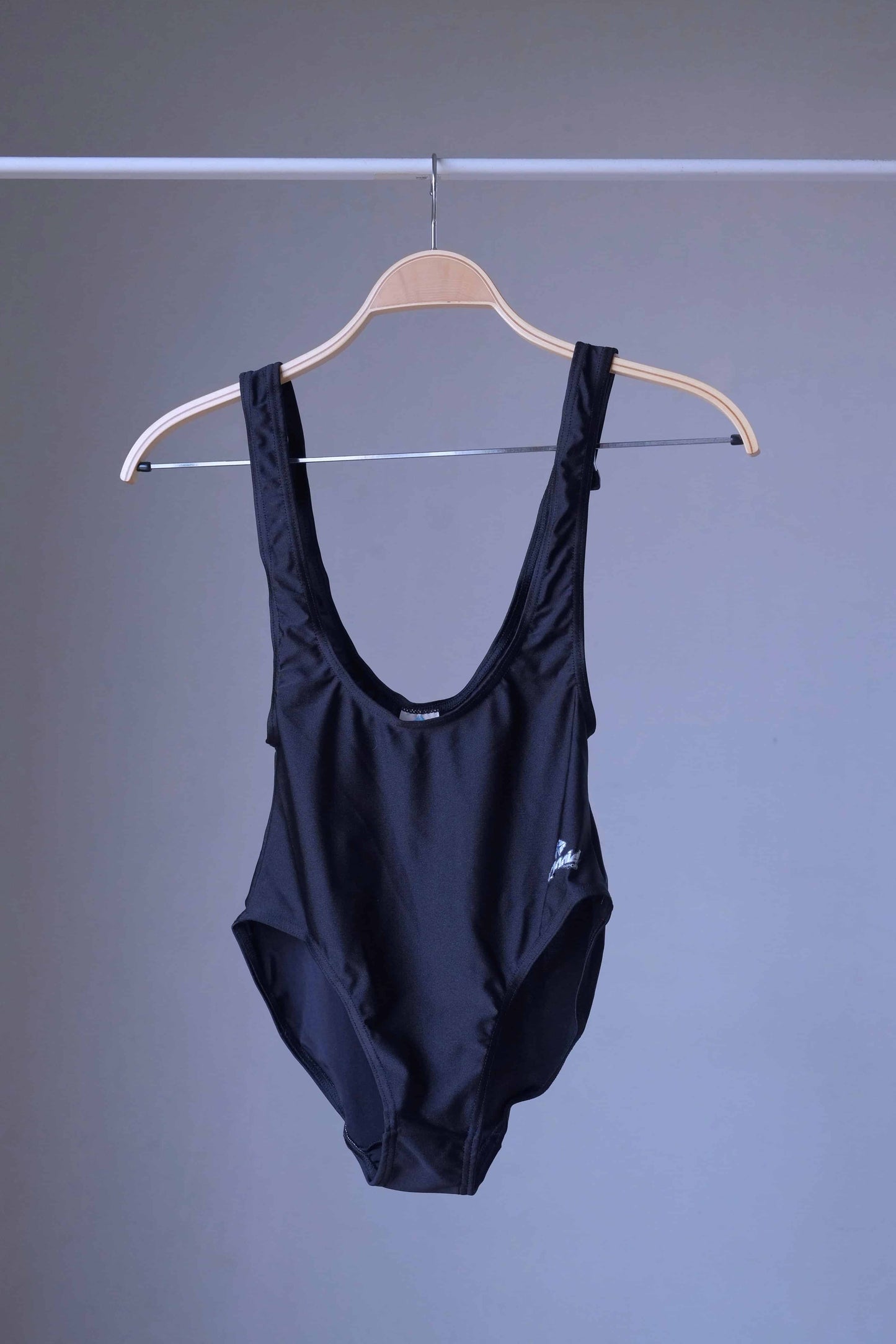 A black, vintage suspender leotard with a deep scoop neck and low back, hanging on a wooden hanger against a gray background.