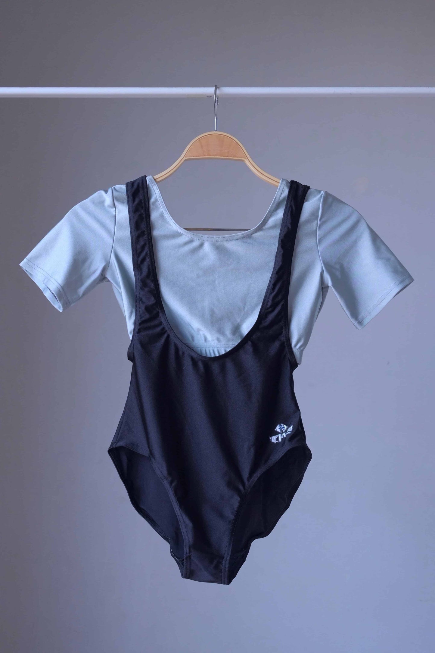 A black, vintage suspender leotard and a silver crop top hanging together on a wooden hanger against a gray background.