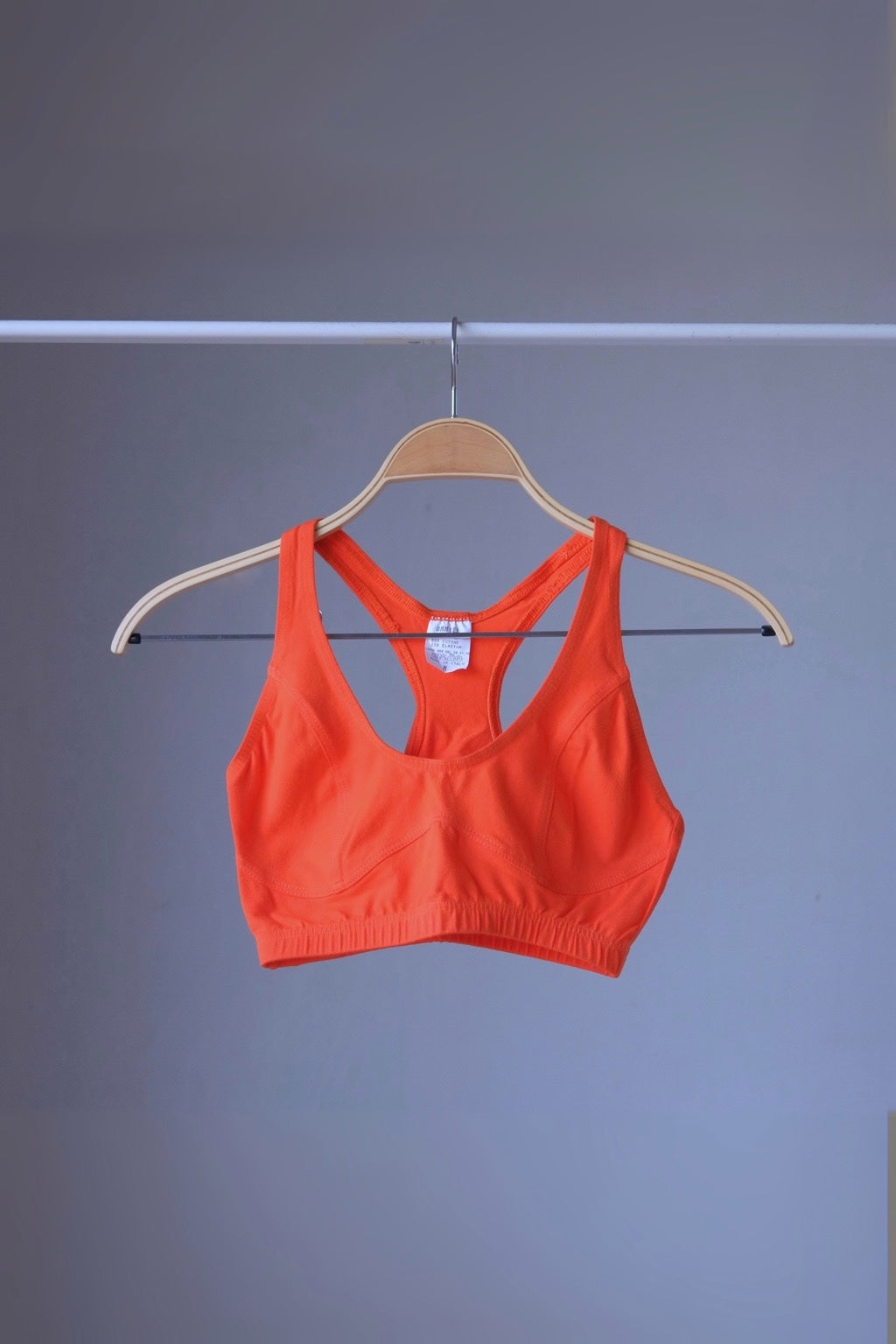 A vibrant orange vintage 90s ANNIEL sports bra with a racerback and elastic underbust band, hanging on a wooden hanger against a gray background.
