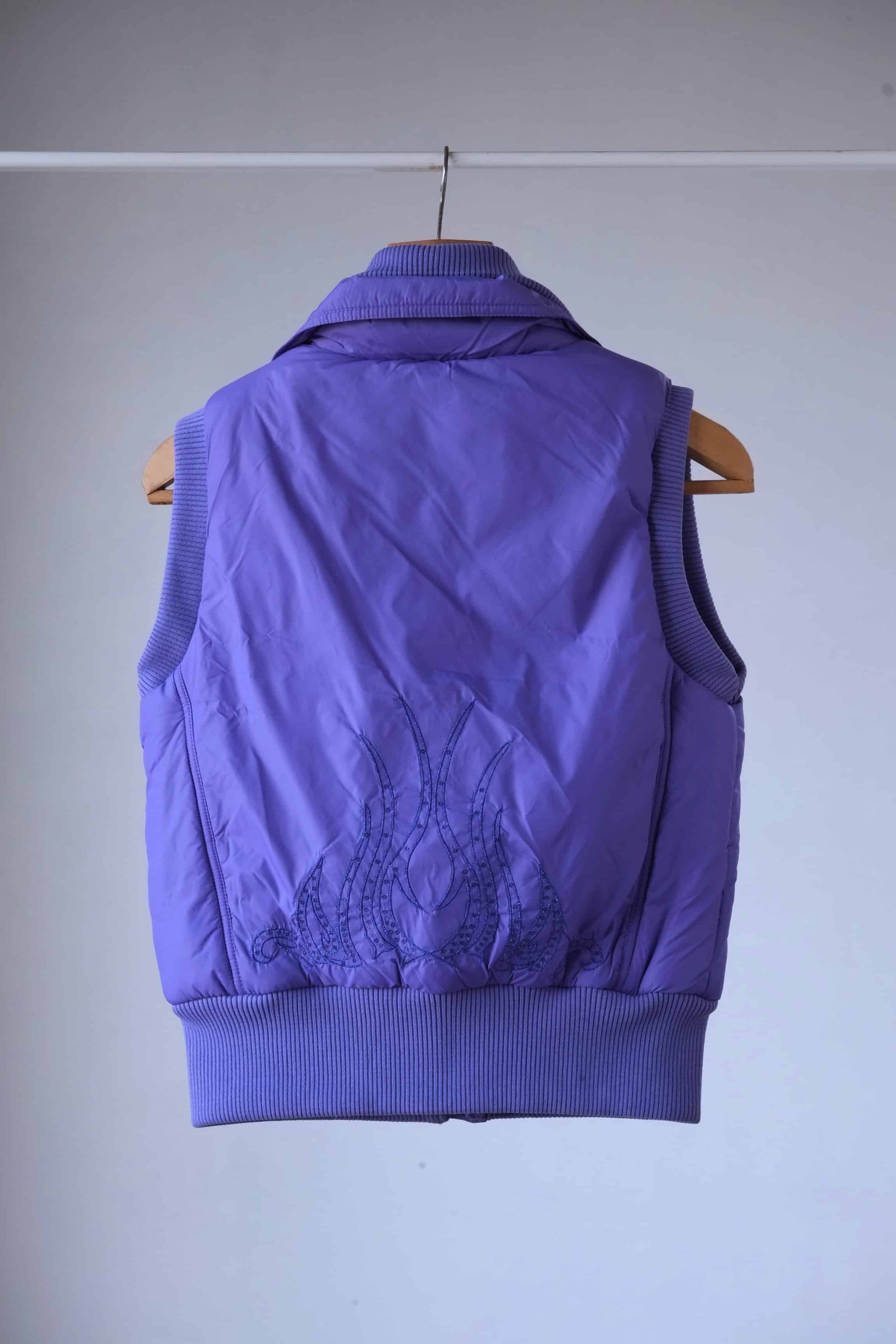 Back view of a purple Y2K-style puffer vest featuring intricate embroidered flame detailing with crystal embellishments. The ribbed hem and collar add a structured yet stylish touch.