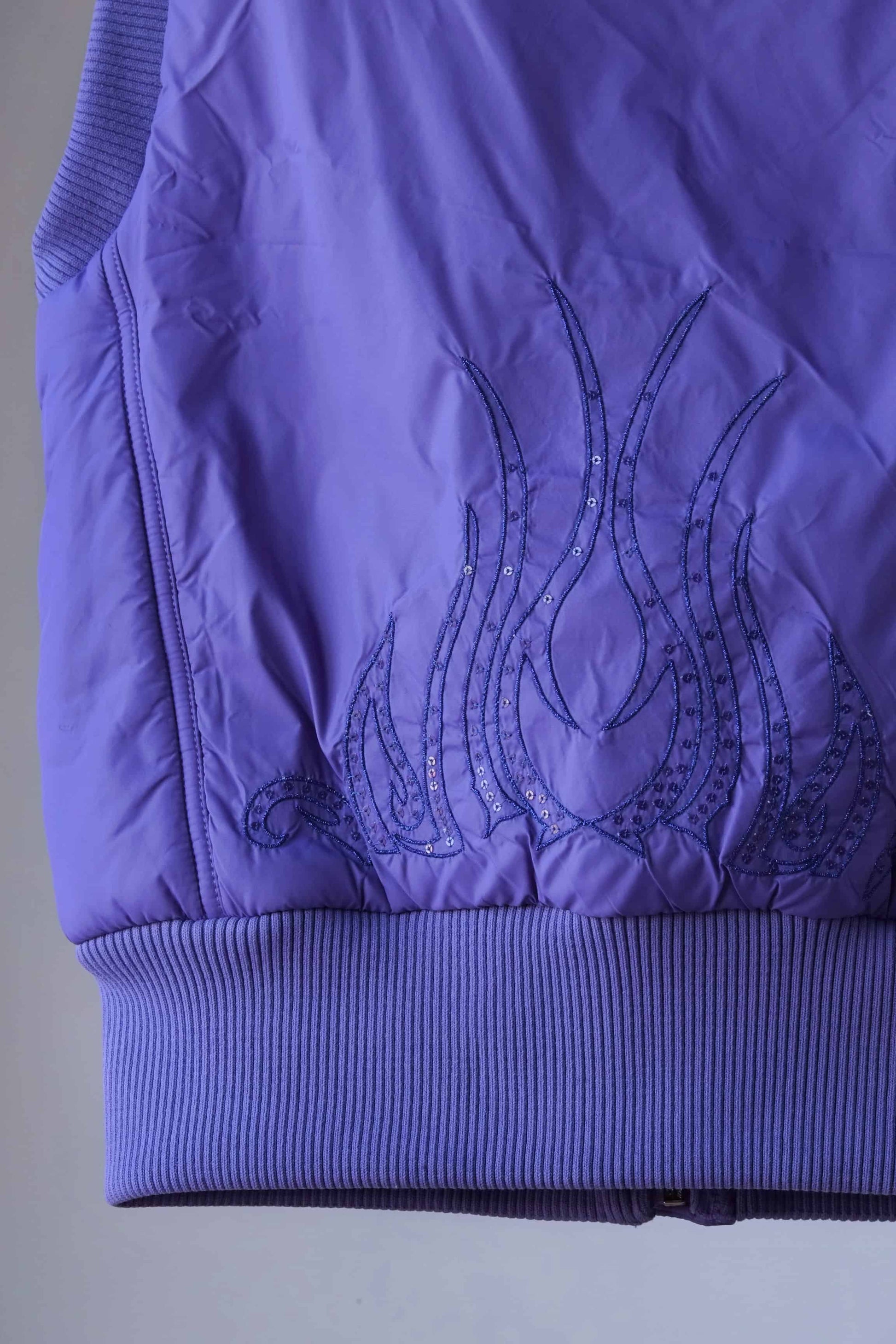 Close-up of the back of a purple Y2K-style puffer vest, showcasing intricate flame embroidery with crystal embellishments. The detailed stitching and ribbed hem add a stylish and textured element to this trendy sleeveless vest.