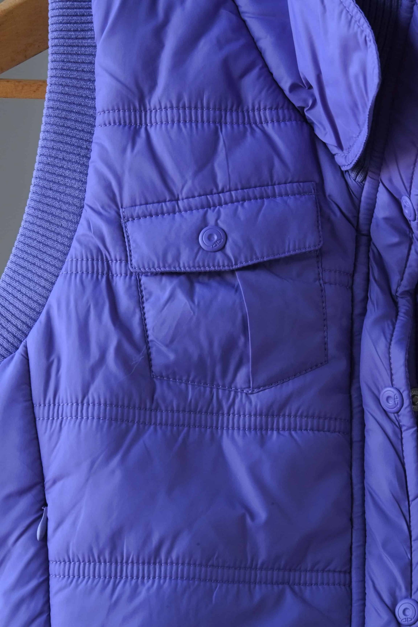 Close-up of a purple Y2K-style puffer vest, highlighting a front snap-button pocket and quilted stitching. The ribbed collar and additional zippered pocket detail are partially visible, showcasing the vest's stylish and functional design.