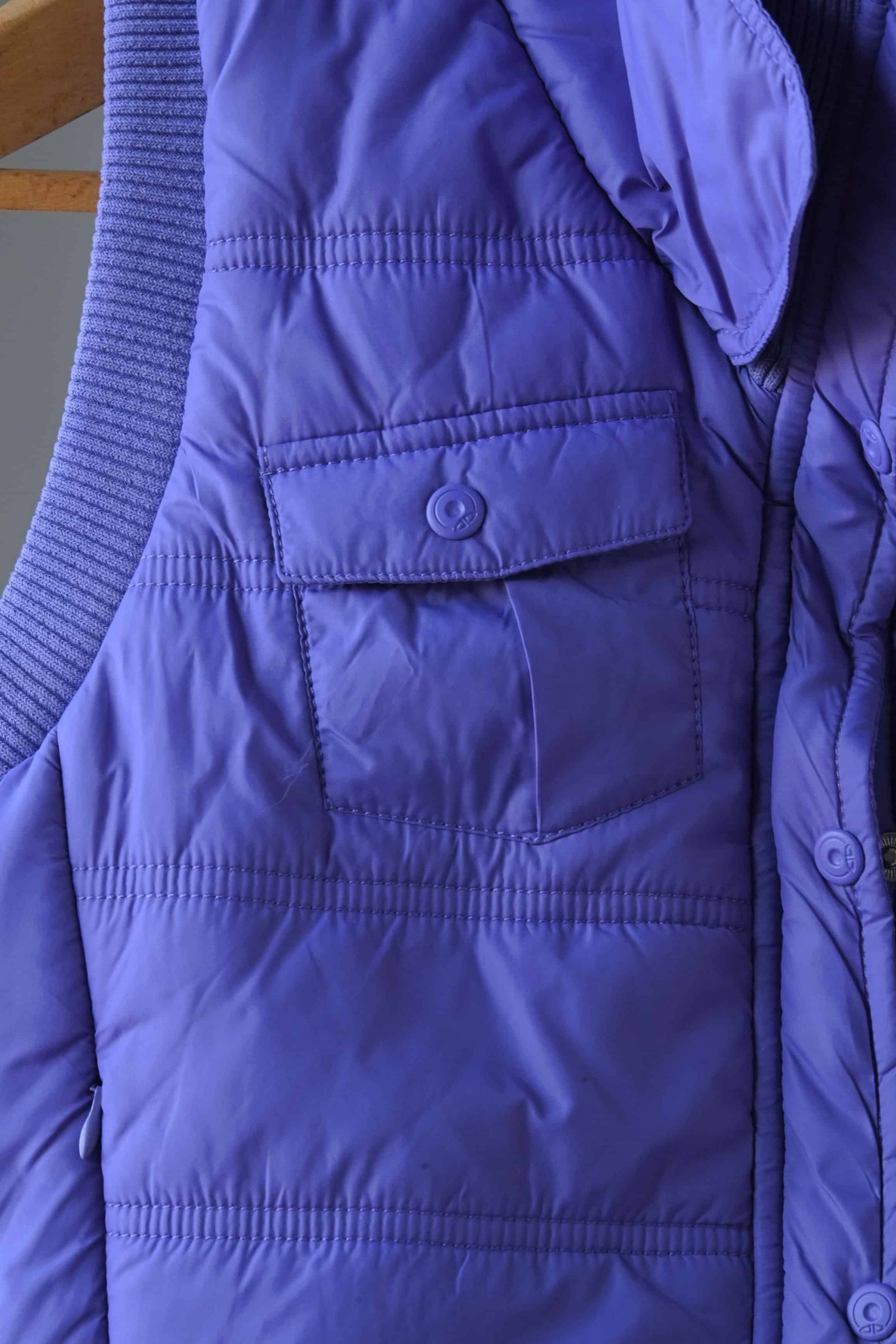 Close-up of a purple Y2K-style puffer vest, highlighting a front snap-button pocket and quilted stitching. The ribbed collar and additional zippered pocket detail are partially visible, showcasing the vest's stylish and functional design.