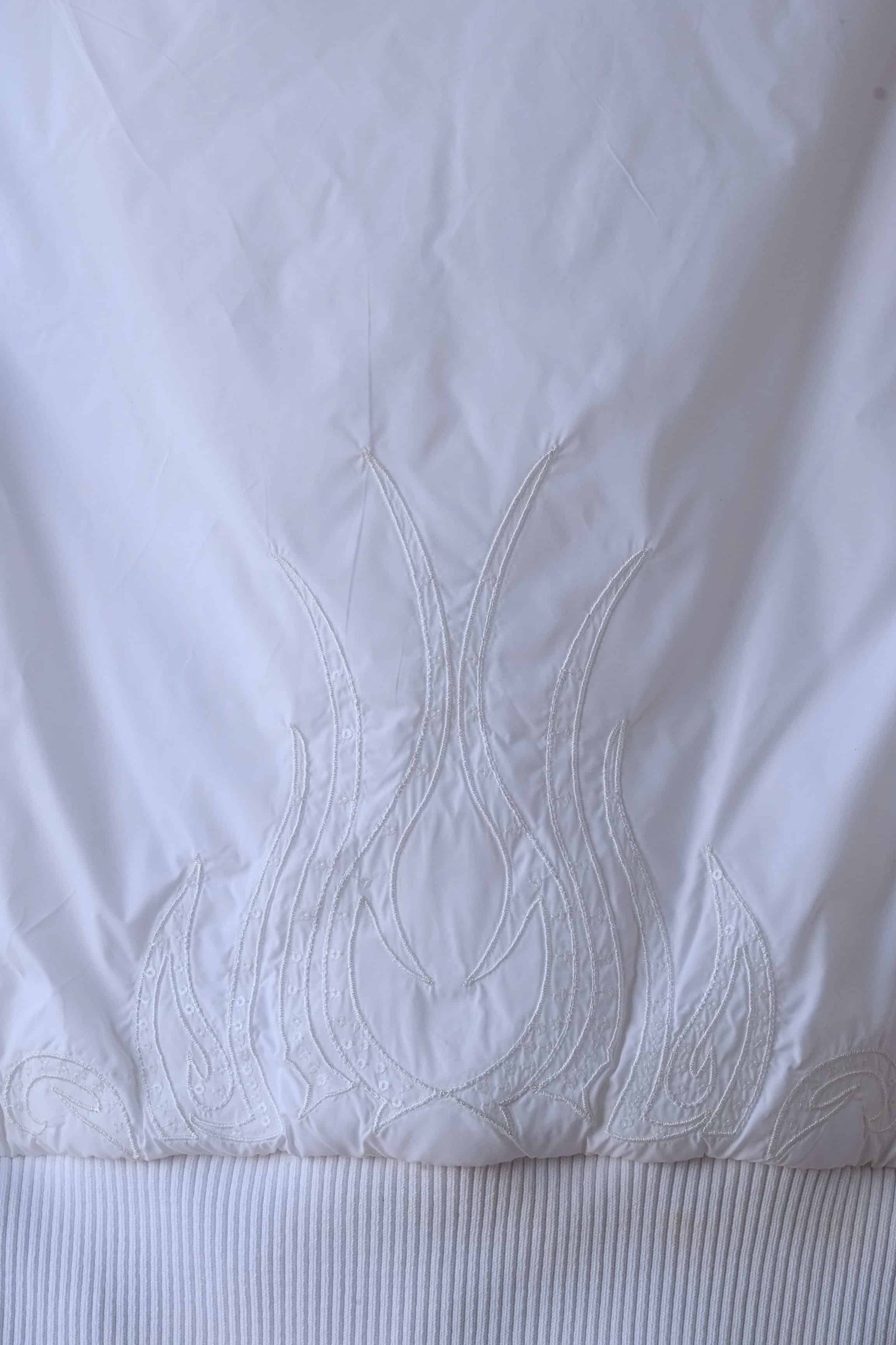 Close-up of the back of a white Y2K-style puffer vest, showcasing intricate flame embroidery with crystal embellishments. The detailed stitching and ribbed hem add a stylish and textured element to this trendy sleeveless vest.