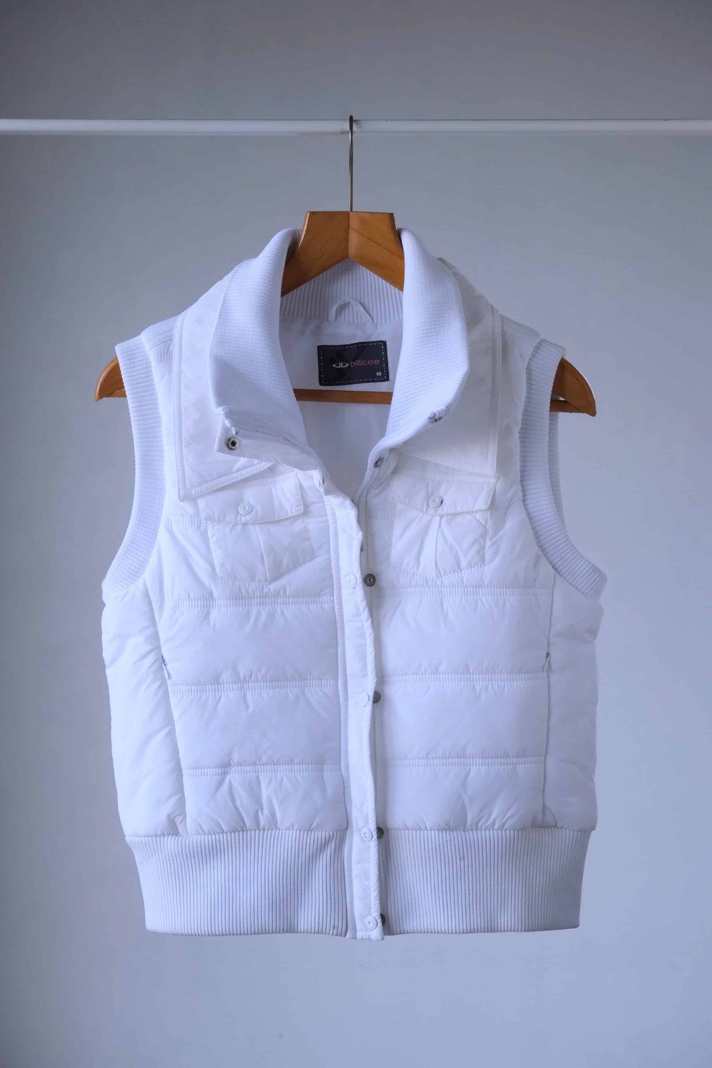A short, sleeveless Y2K white puffer vest. It has a high collar and a zipper closure. The vest is hanging on a wooden hanger against a light gray background.