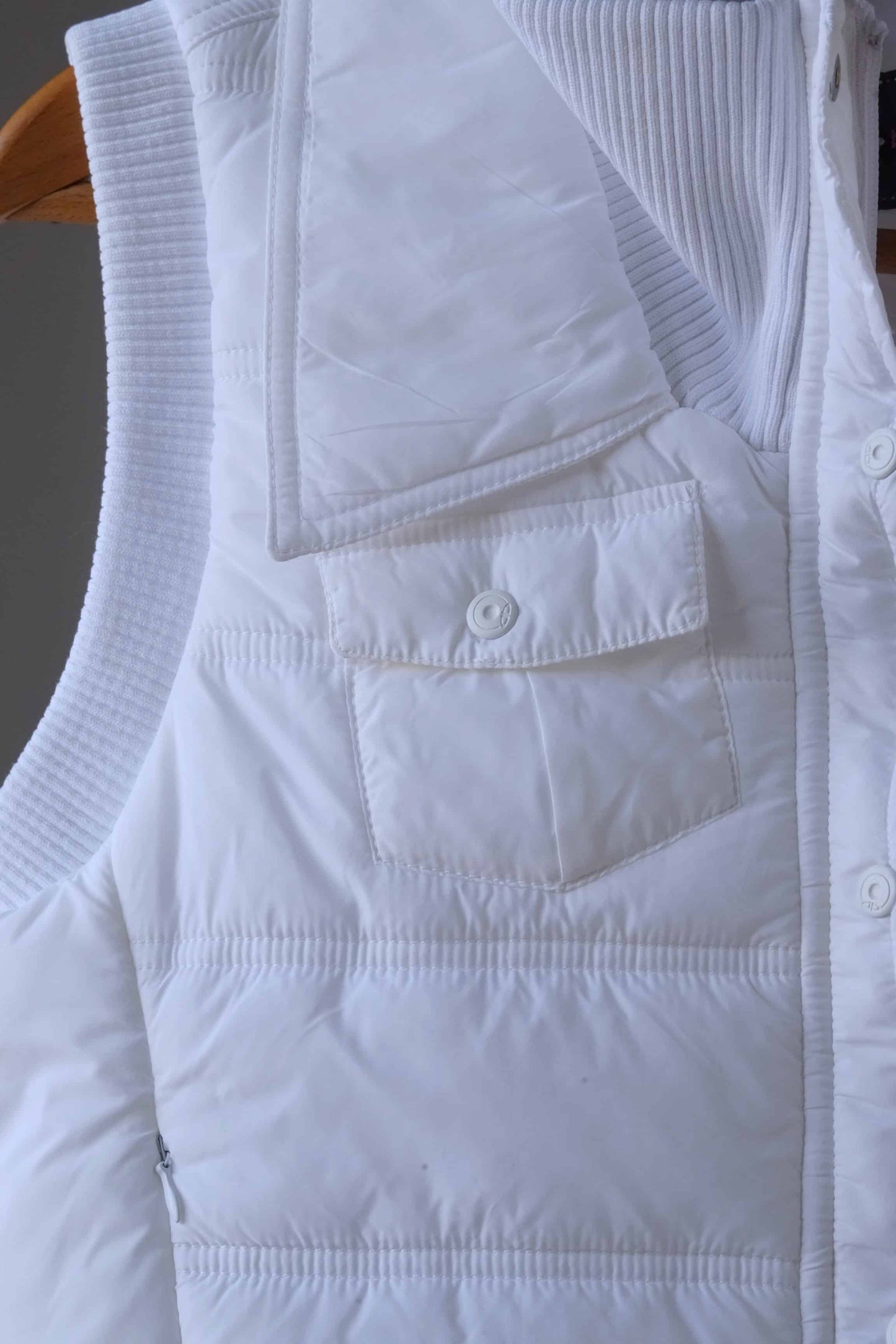 Close-up of a white Y2K-style puffer vest, highlighting a front snap-button pocket and quilted stitching. The ribbed collar and additional zippered pocket detail are partially visible, showcasing the vest's stylish and functional design.