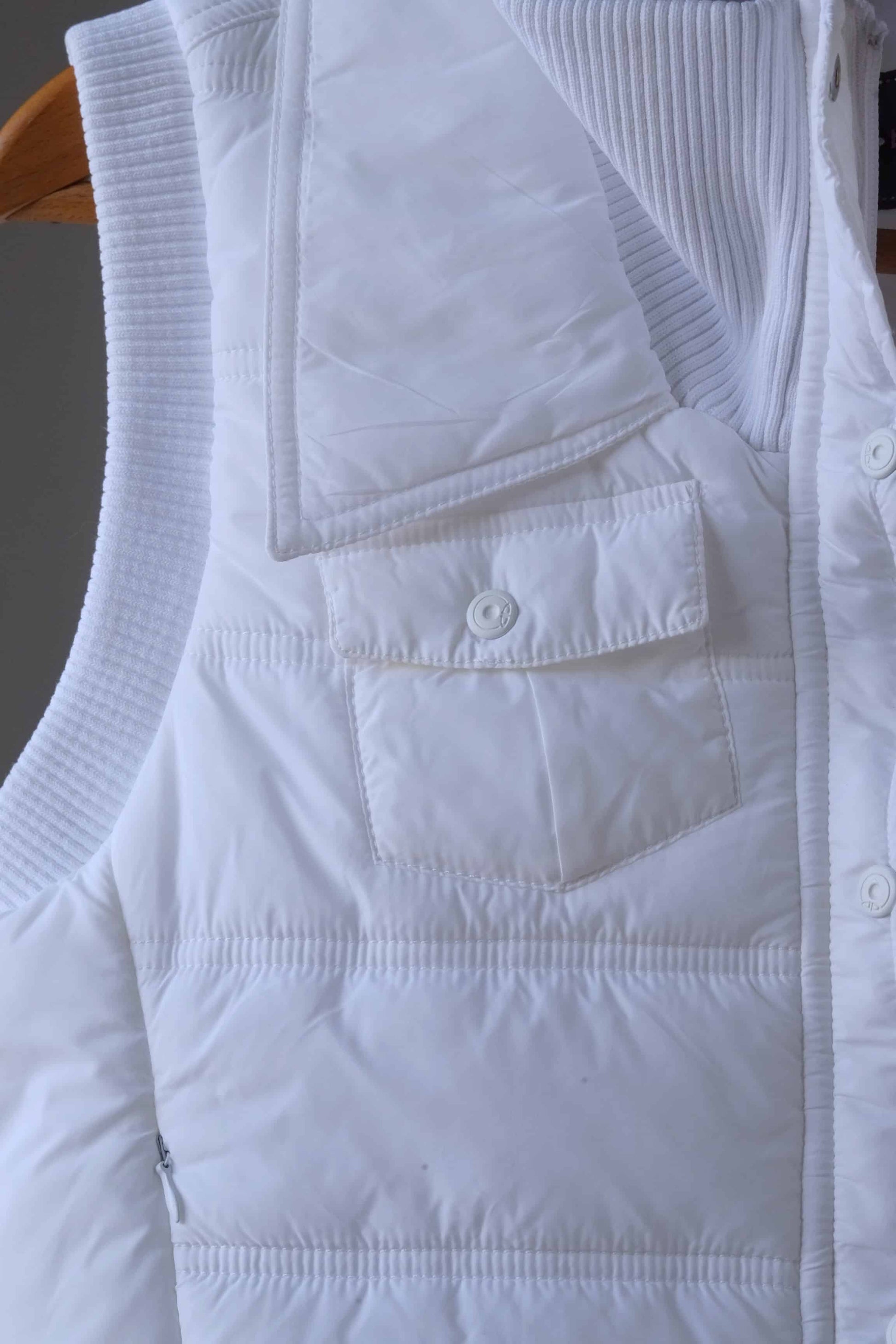 Close-up of a white Y2K-style puffer vest, highlighting a front snap-button pocket and quilted stitching. The ribbed collar and additional zippered pocket detail are partially visible, showcasing the vest's stylish and functional design.