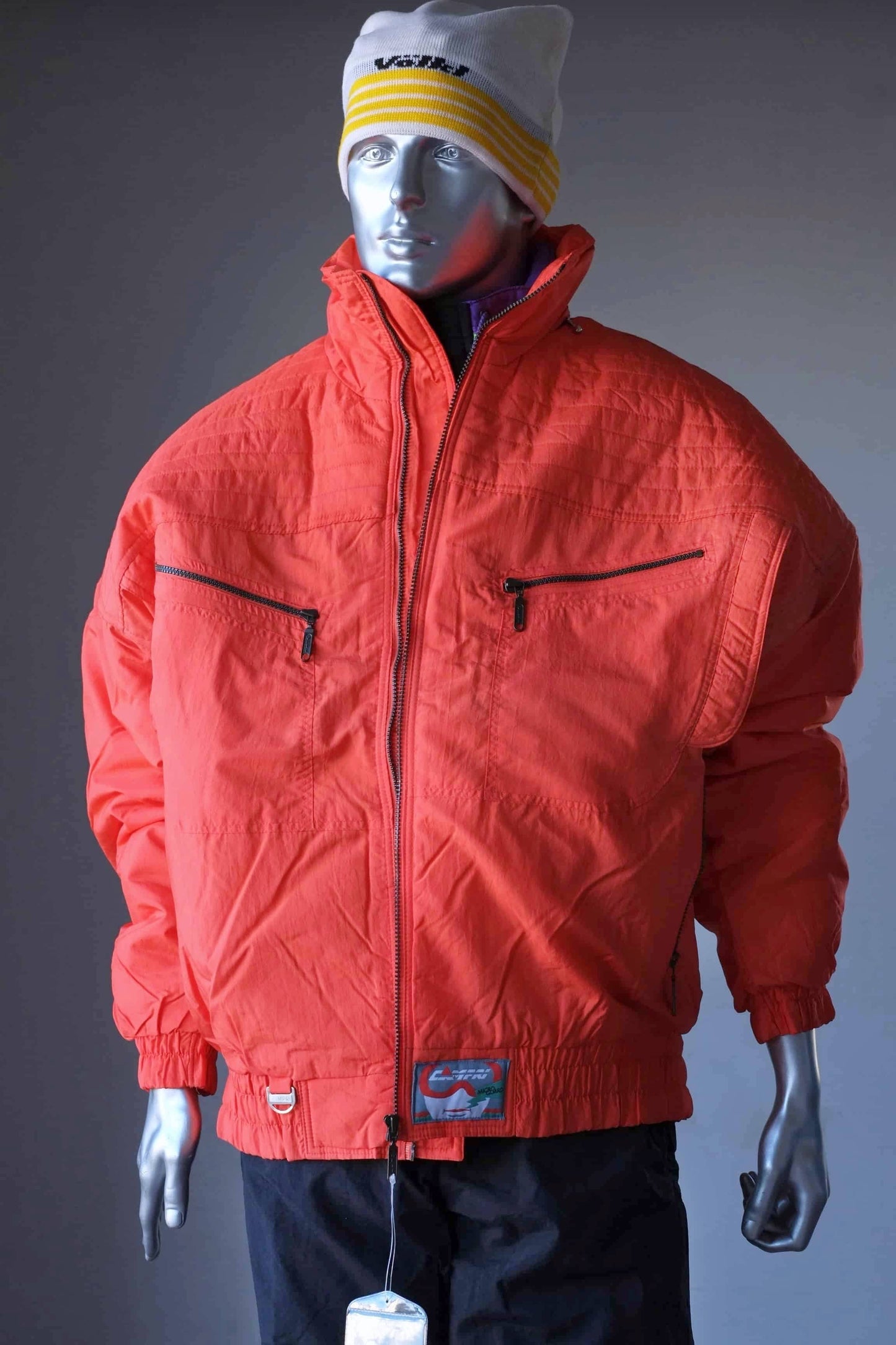 A mannequin wearing A bright orange vintage ski jacket hanging The jacket has a puffy design with multiple pockets and a drawstring waist. It features a retro-style logo with the text "Campri" and "Microguard." The mannequin is also wearing a yellow and white wool beanie.