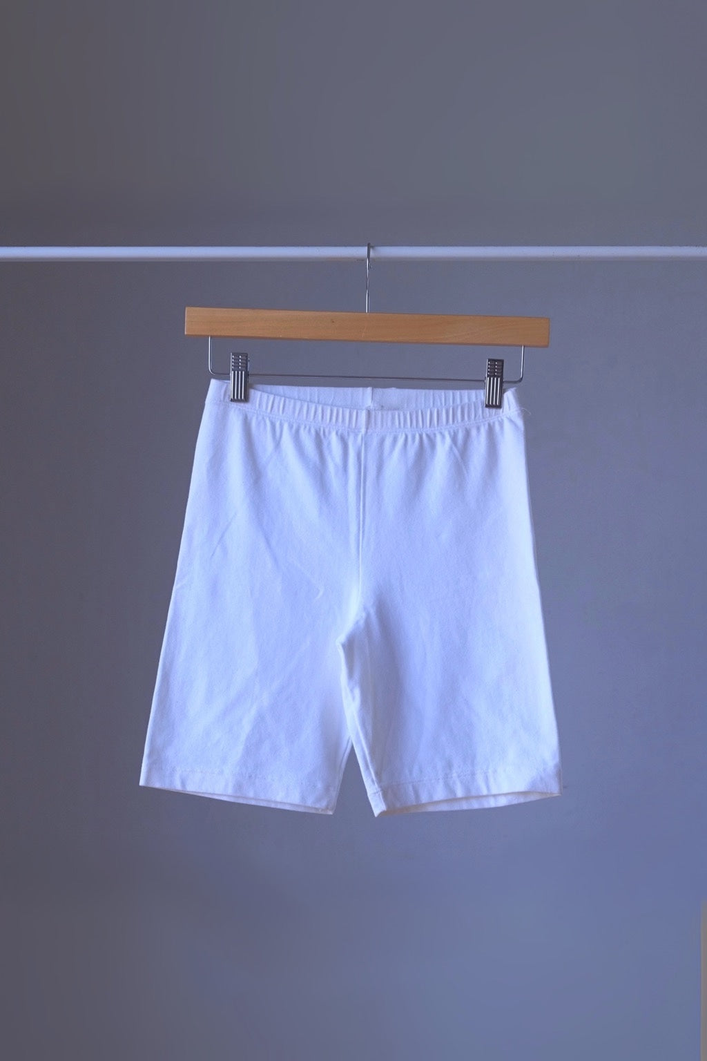 A pair of white vintage 90s DANSKIN bike shorts hanging on a wooden hanger against a gray background.