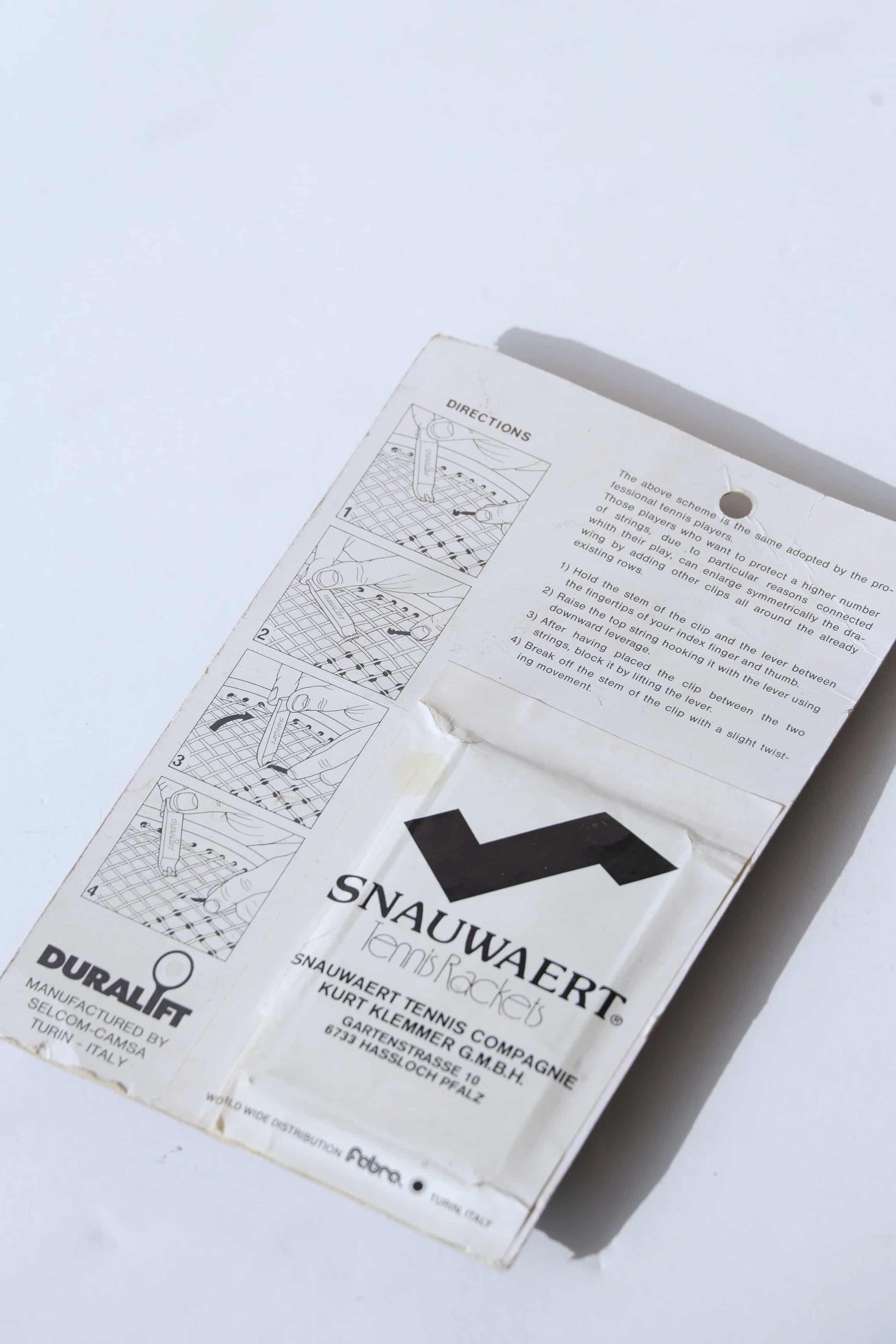 Close up of the Backside of a vintage Duralift Micrograph Tennis String Saver Kit. It is white with black text. The word SNAUWAERT is visible with a stylized S which looks like the logo of the brand.