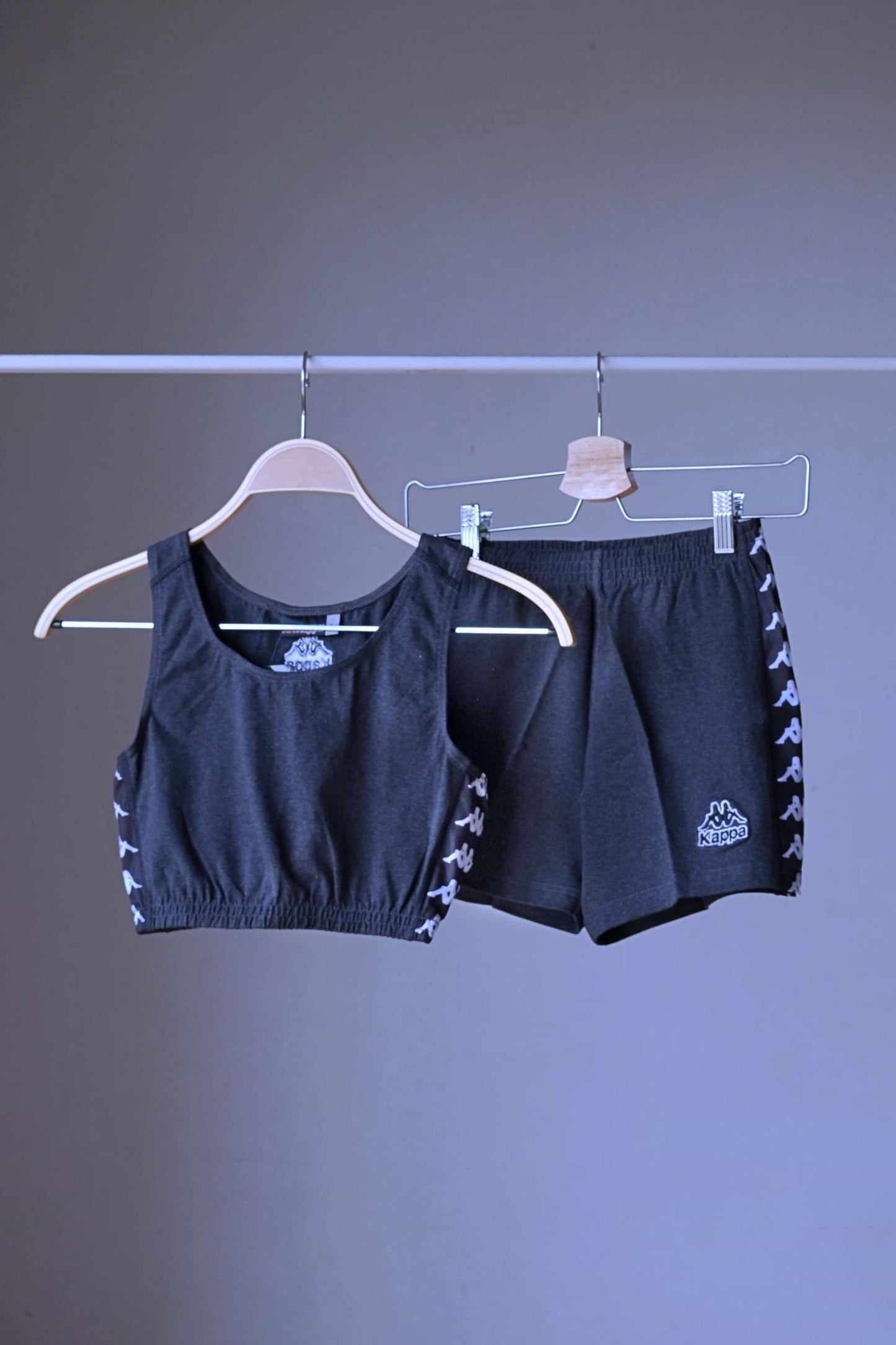 Front view of a charcoal grey KAPPA vintage women's athletic crop top and shorts set on a hanger, with the shorts hanging behind the top, displaying the white logo taping on both garments.