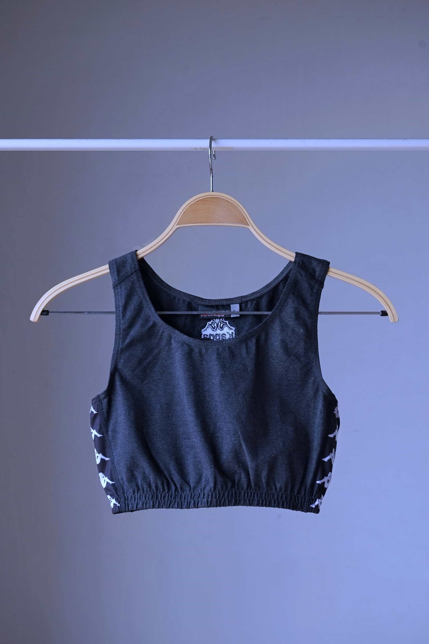 Front view of a charcoal grey KAPPA vintage women's athletic crop top on a wooden hanger, showing the KAPPA logo on the front and white logo taping along the sides.