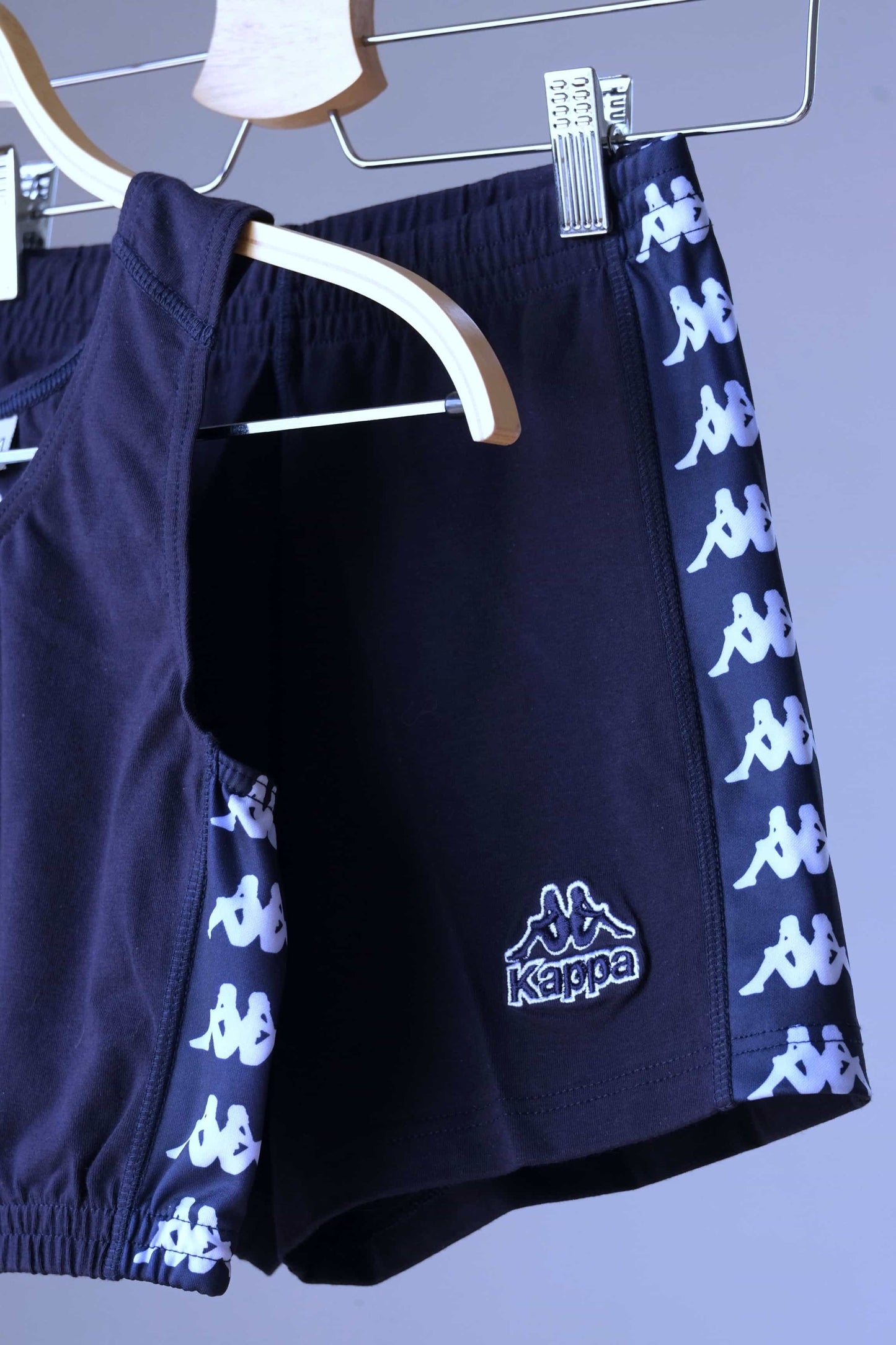 Close-up view of a navy blue KAPPA vintage women's athletic crop top and shorts set on hangers, highlighting the white KAPPA logo taping on the sides of both garments and the KAPPA logo on the shorts.