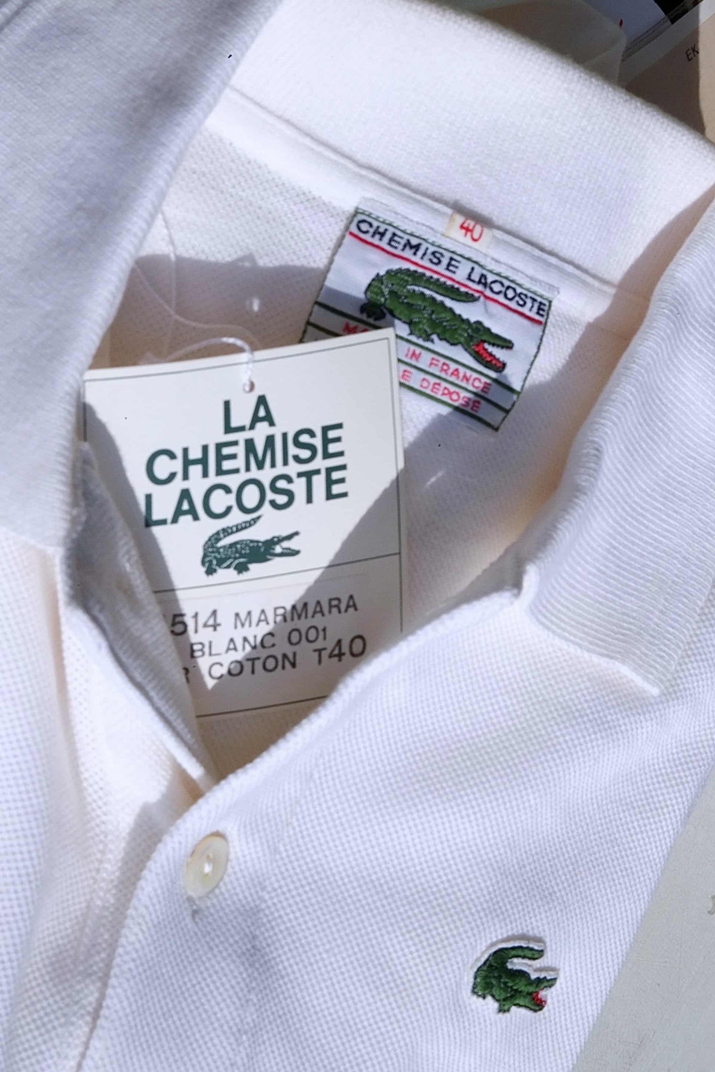 Close-up of a white Lacoste polo shirt collar tag. The tag displays the Lacoste crocodile logo, size, fabric details, and "La Chemise Lacoste" text in French.