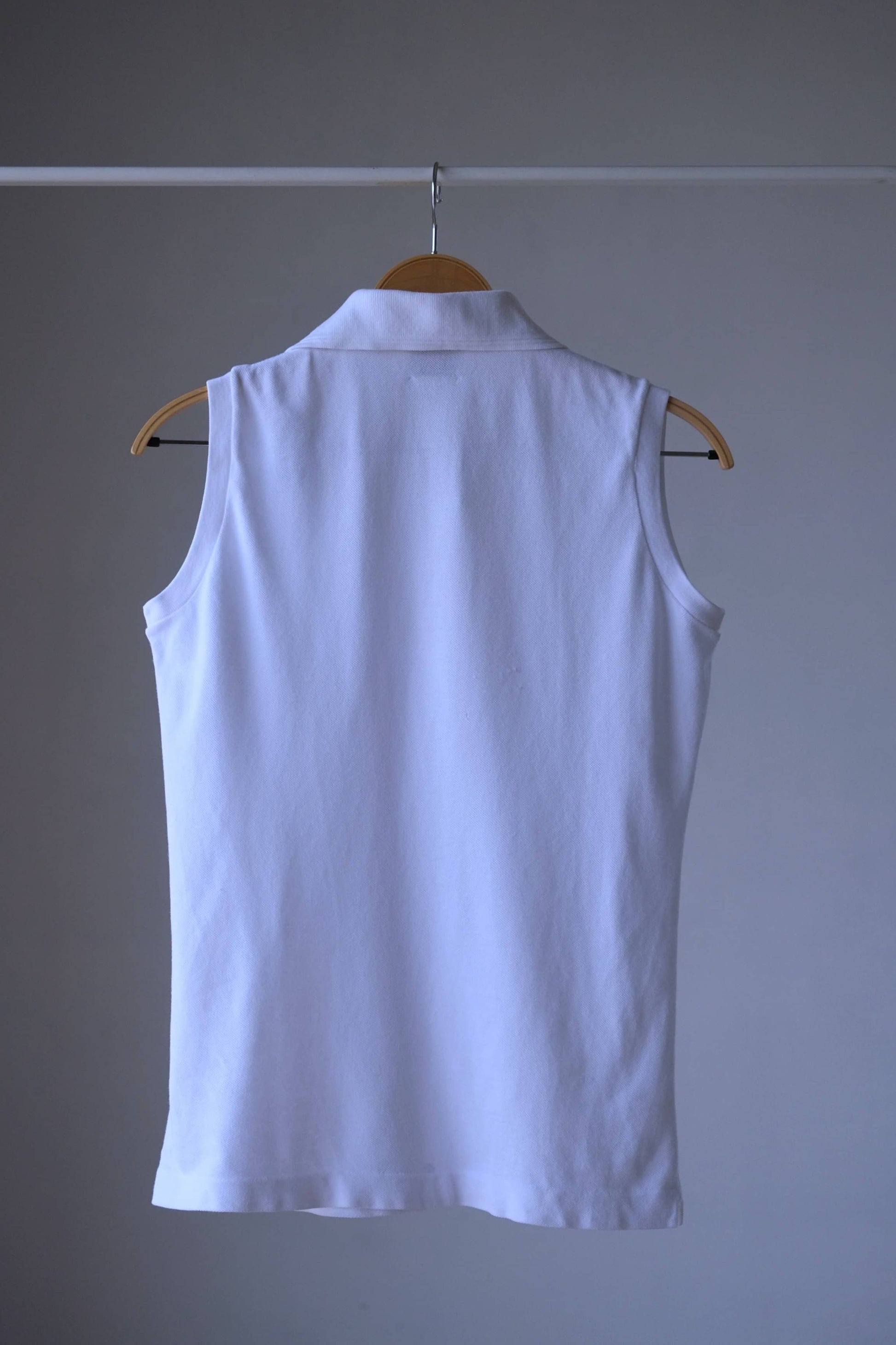 Back view of a white sleeveless Lacoste polo shirt hanging on a wooden hanger, showing the collar.