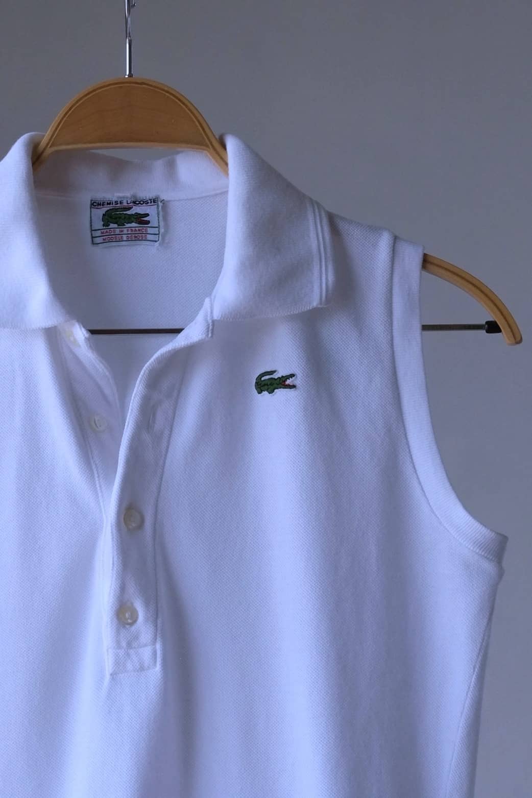 Close-up of a white sleeveless Lacoste polo shirt hanging on a wooden hanger. The shirt features a collar, button placket,  and the iconic Lacoste crocodile logo embroidered on the right chest. The label on the inside of the collar reads "CHEMISE LACOSTE".