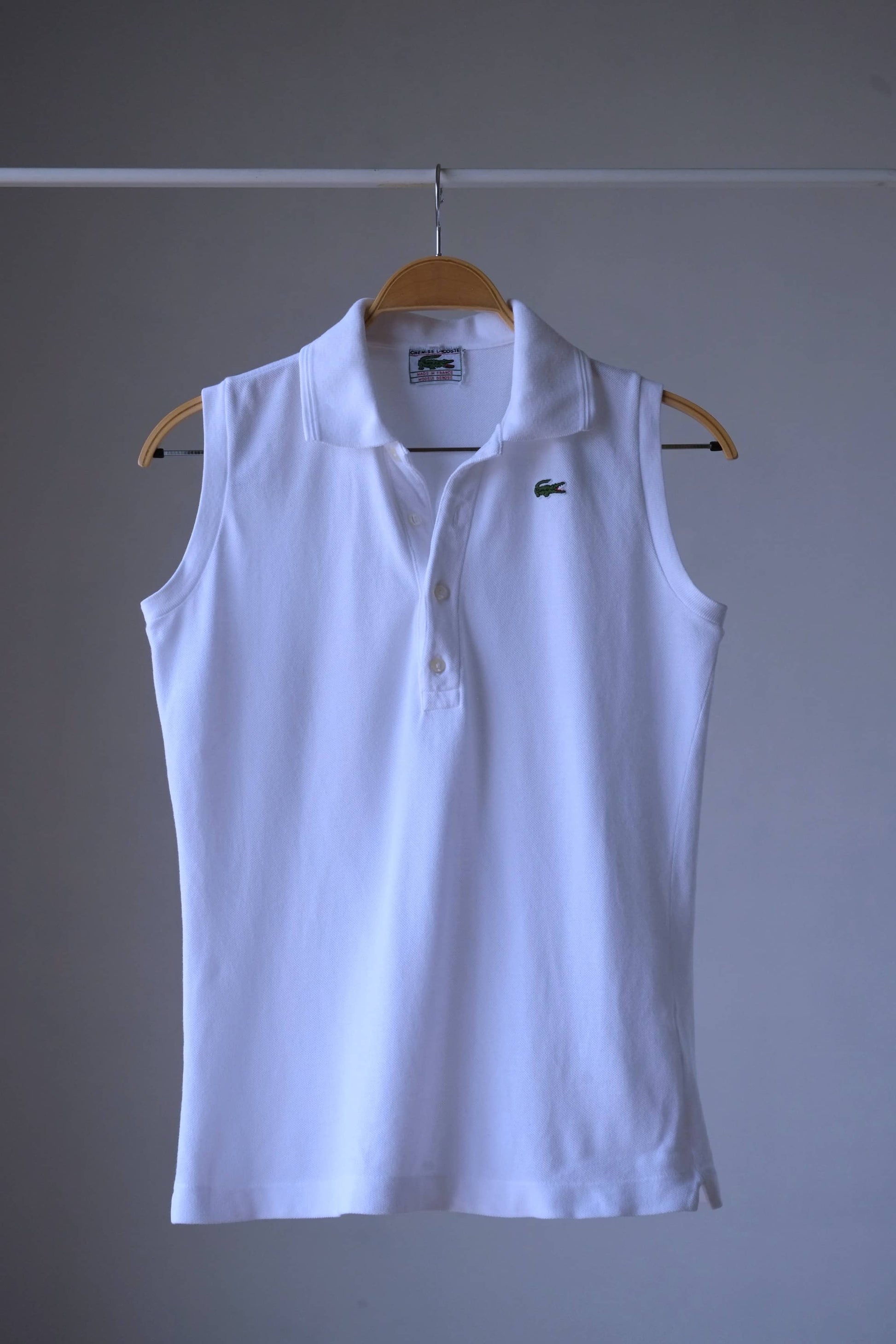 White sleeveless Lacoste polo shirt with a collar and button placket, hanging on a wooden hanger against a neutral background. The iconic Lacoste crocodile logo is embroidered on the left chest.