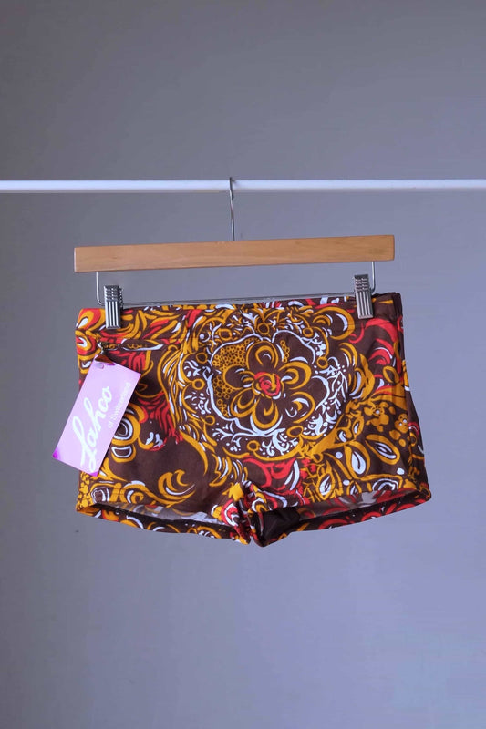 Front view of LAHCO vintage 1960s swim trunks on a hanger. The trunks are brown with intricate gold, yellow, and red psychedelic floral patterns and feature a side zip pocket and a Lahco tag.