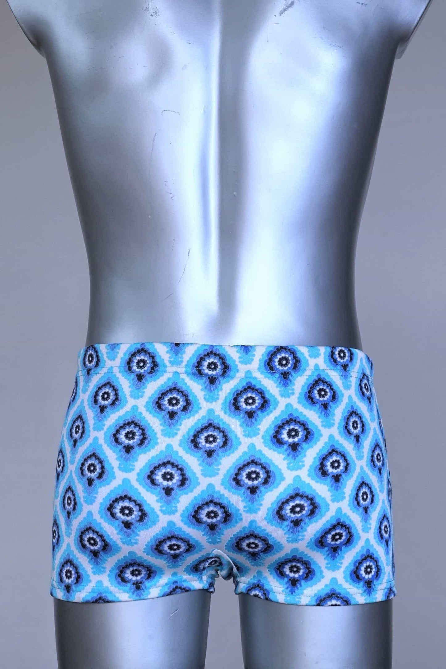 Back view of vintage 60s Lahco swim trunks in icy blue with a repeating dark blue and white geometric medallion pattern, featuring a zip pocket and brand tag.