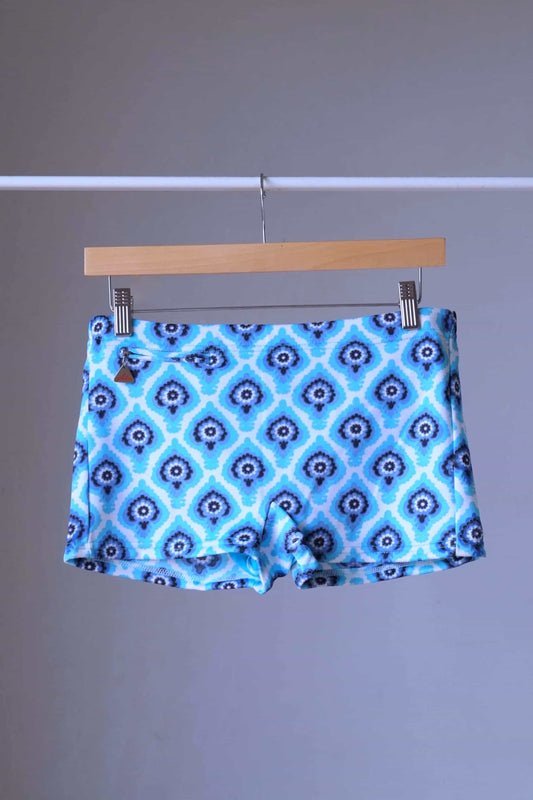 Vintage 60s Lahco swim trunks in blue with a geometric medallion print, shown on a wooden hanger, featuring a front drawstring and zip pocket.