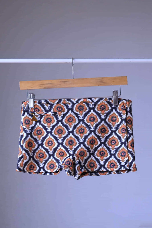 Vintage 60s Lahco swim trunks in brown with a geometric medallion print, displayed on a wooden hanger, featuring a front drawstring and zip pocket.