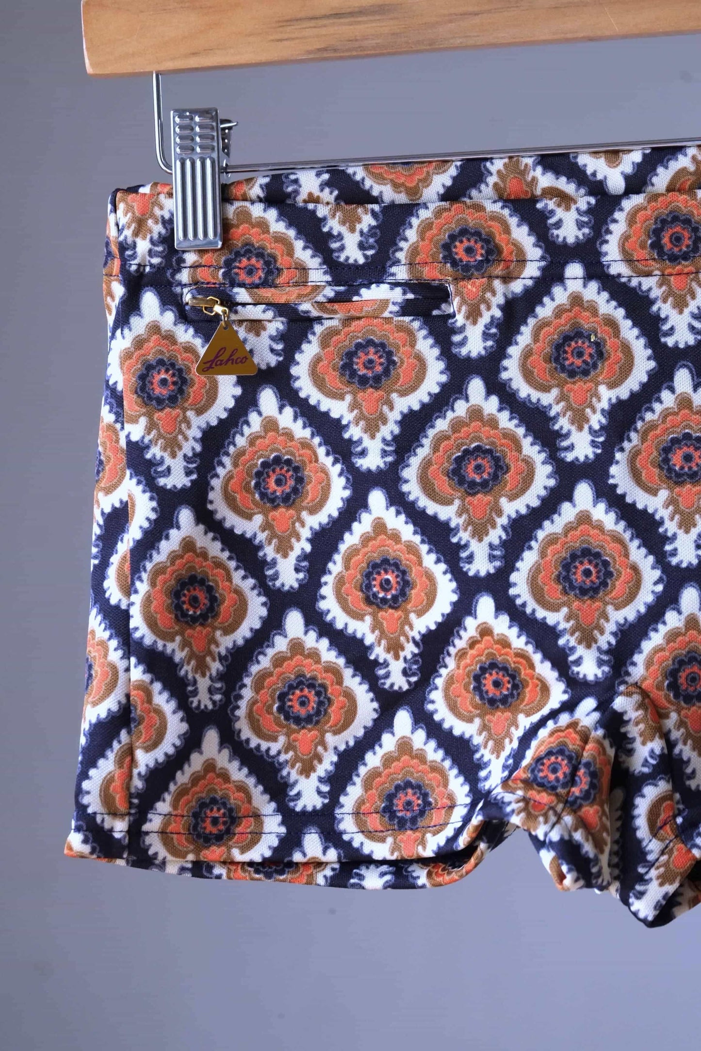 Close view of a Vintage 60s Lahco swim trunks in brown with a geometric medallion print, displayed on a wooden hanger, featuring a front drawstring and zip pocket.