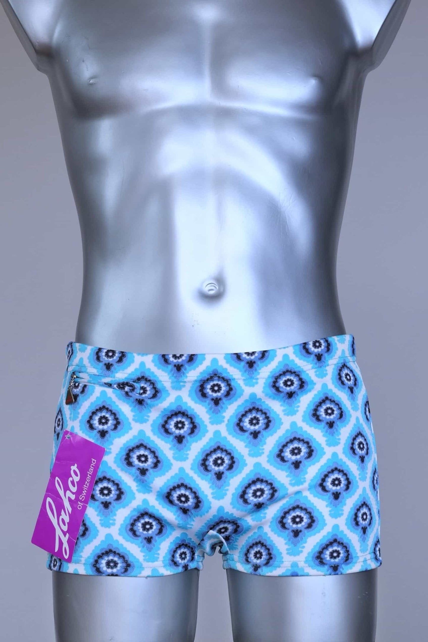 Front view of vintage 60s Lahco swim trunks in icy blue with a repeating dark blue and white geometric medallion pattern, featuring a zip pocket and brand tag.