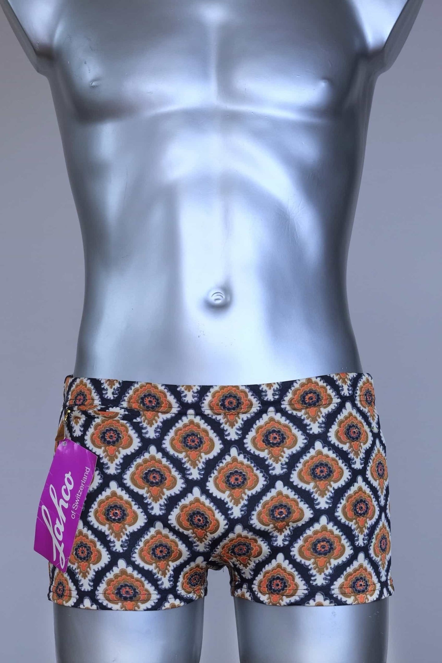 Front view of vintage 60s Lahco swim trunks in navy with an allover brown, orange, and cream geometric medallion print, featuring a zip pocket and original brand tag.