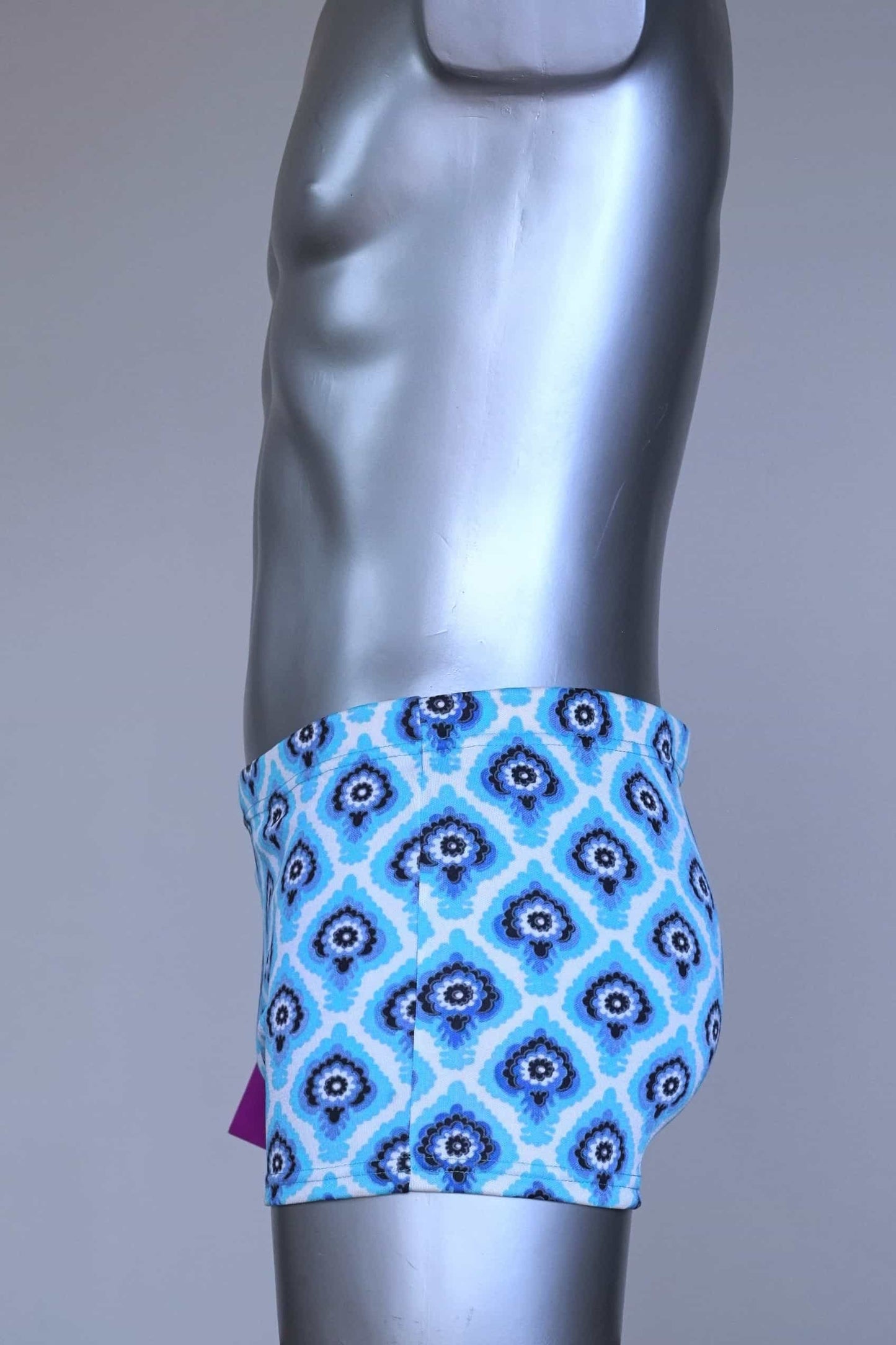Side view of vintage 60s Lahco swim trunks in icy blue with a repeating dark blue and white geometric medallion pattern, featuring a zip pocket and brand tag.