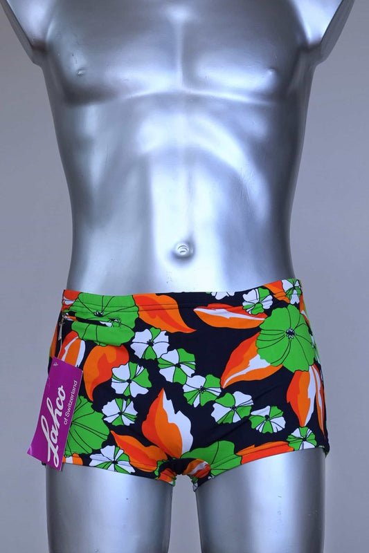 Front view of LAHCO vintage 1960s men's swim trunks on a silver mannequin. The trunks are black with a vibrant green, orange, and white floral pattern. Shown here with their original tag.