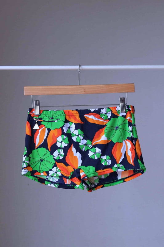 Front view of LAHCO vintage 1960s men's swim trunks on a wooden hanger. The trunks are black with a vibrant green, orange, and white floral pattern.