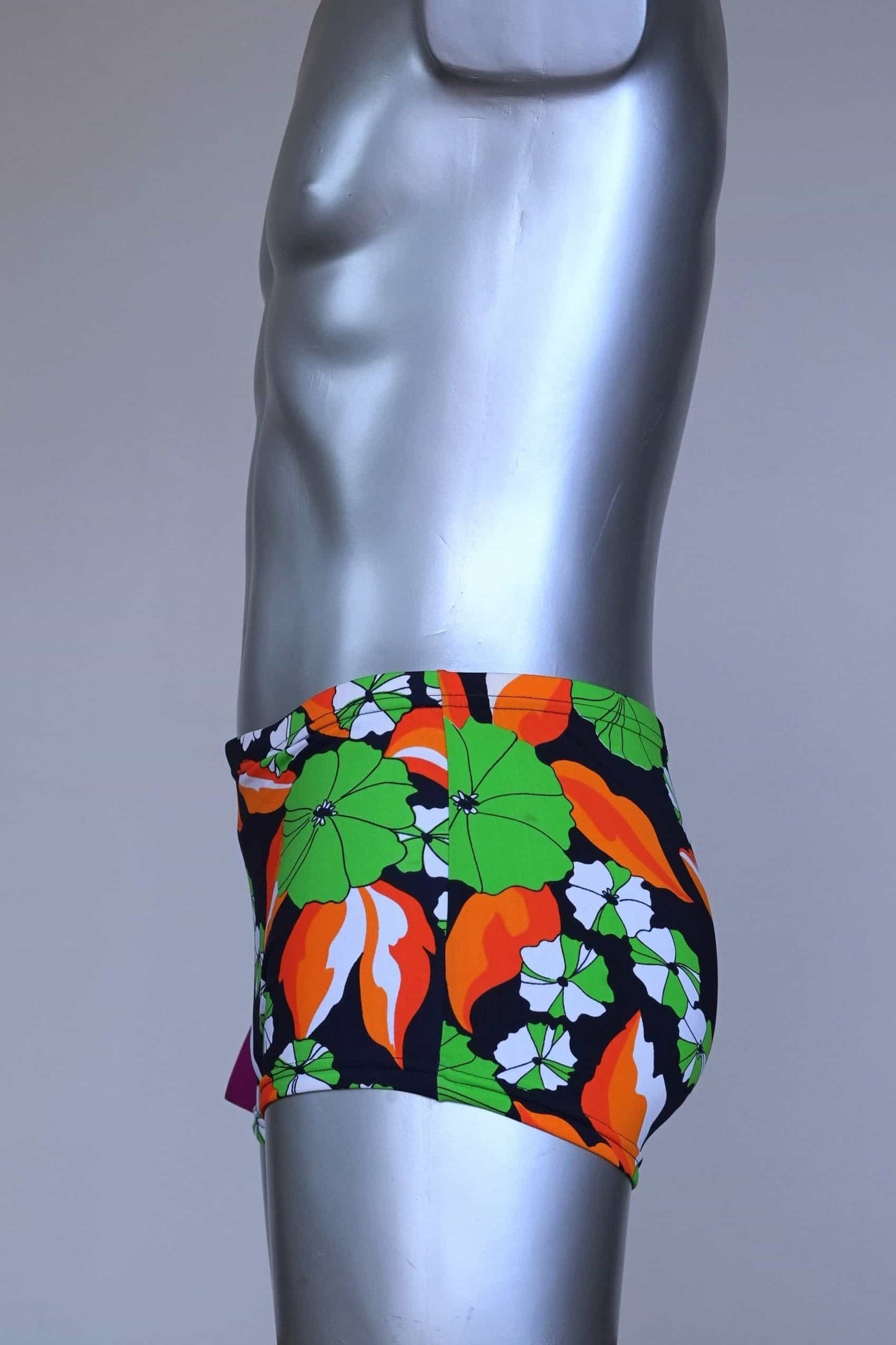 Side view of LAHCO vintage 1960s men's swim trunks on a silver mannequin. The trunks are black with a vibrant green, orange, and white floral pattern.