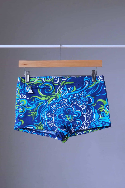Front view of LAHCO vintage 1960s swim trunks on a hanger. The trunks are brown with intricate blue, green, and white psychedelic floral patterns and feature a side zip pocket and a Lahco tag.