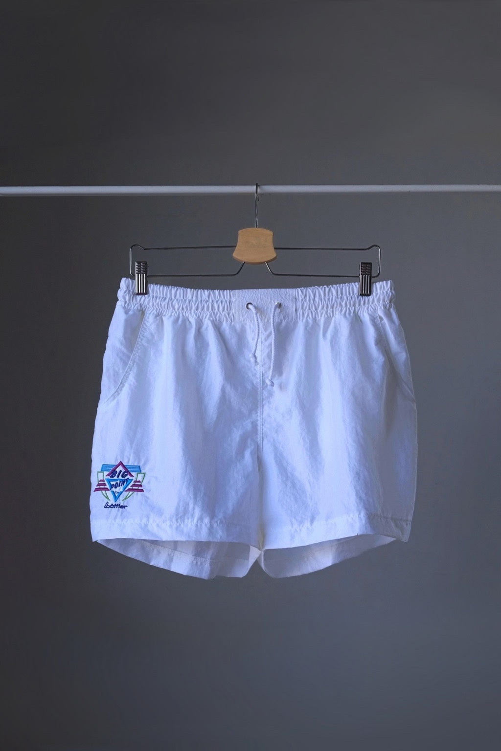 Front view of a pair of white vintage Löffler shorts with a small logo on the left leg, hanging on a hanger.