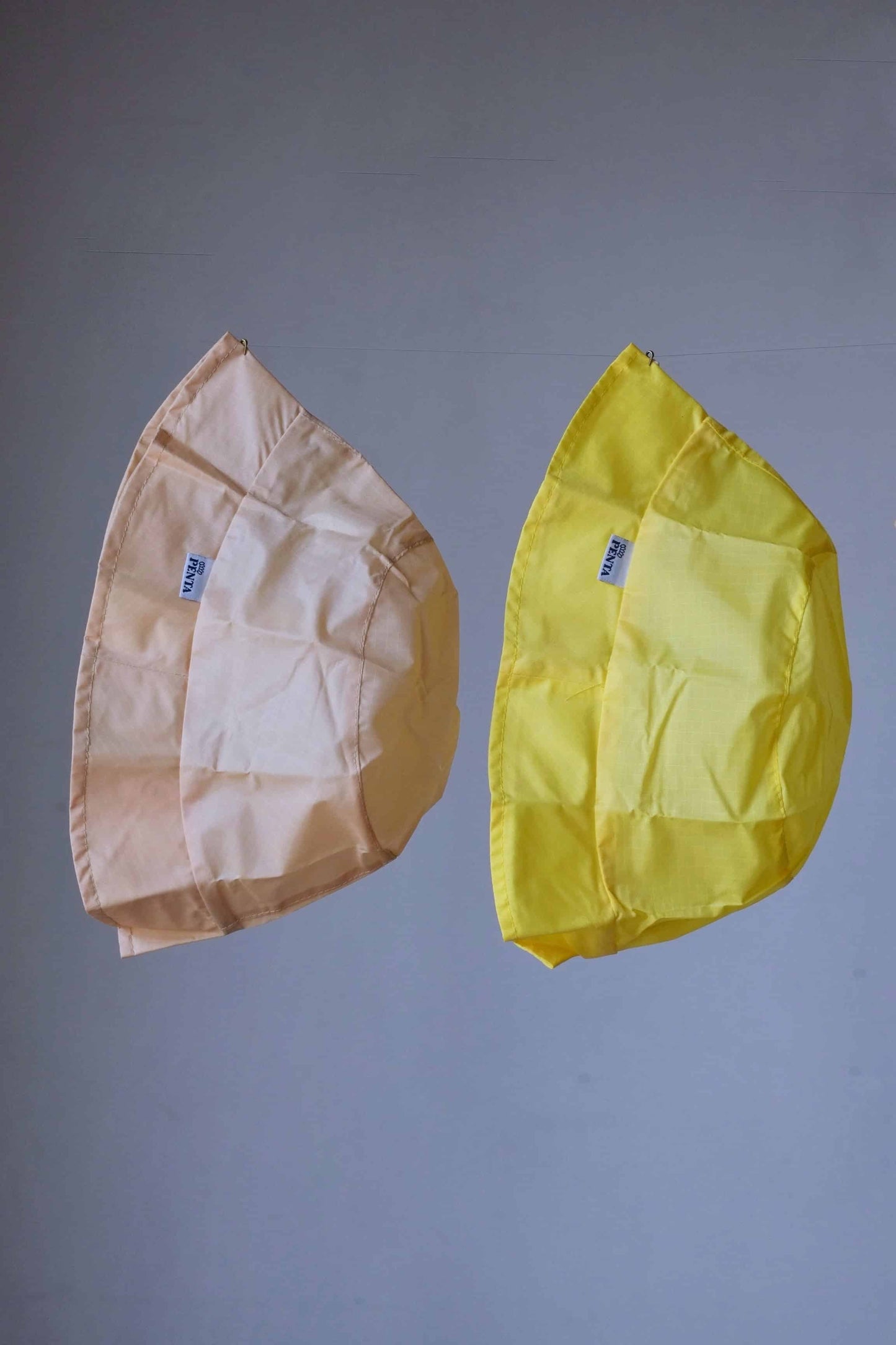 Two rainproof bucket hats, a yellow and a beige one, hanging in front of a grey background