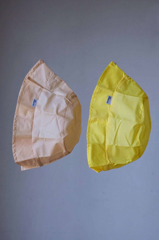 Two rainproof bucket hats, a yellow and a beige one, hanging in front of a grey background