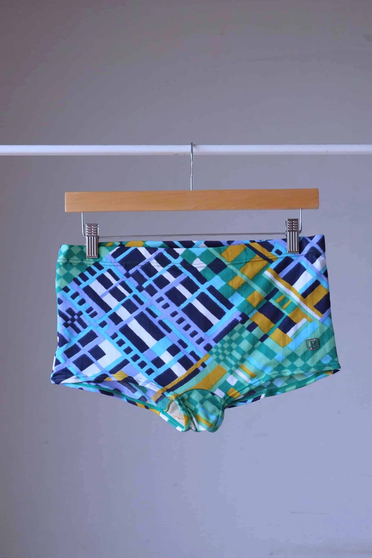 Ribana vintage 70s geometric swim trunks in blue and green with abstract check pattern, hanging on wooden hanger.
