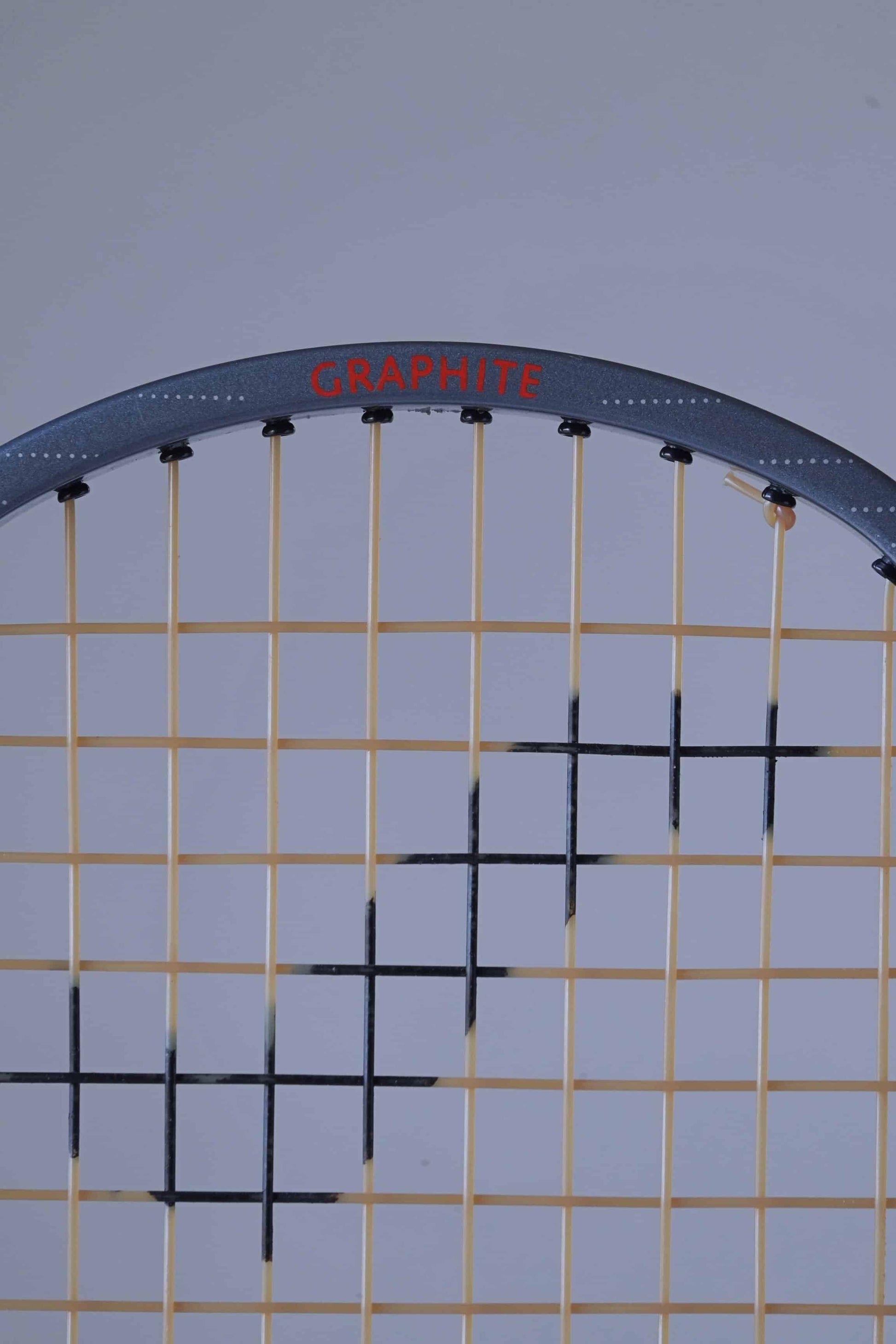 Close-up of the top section of a Snauwaert Hi-Ten 30 tennis racket, showing the word 'GRAPHITE' in red on the grey frame, with yellow strings and a black patterned string design.