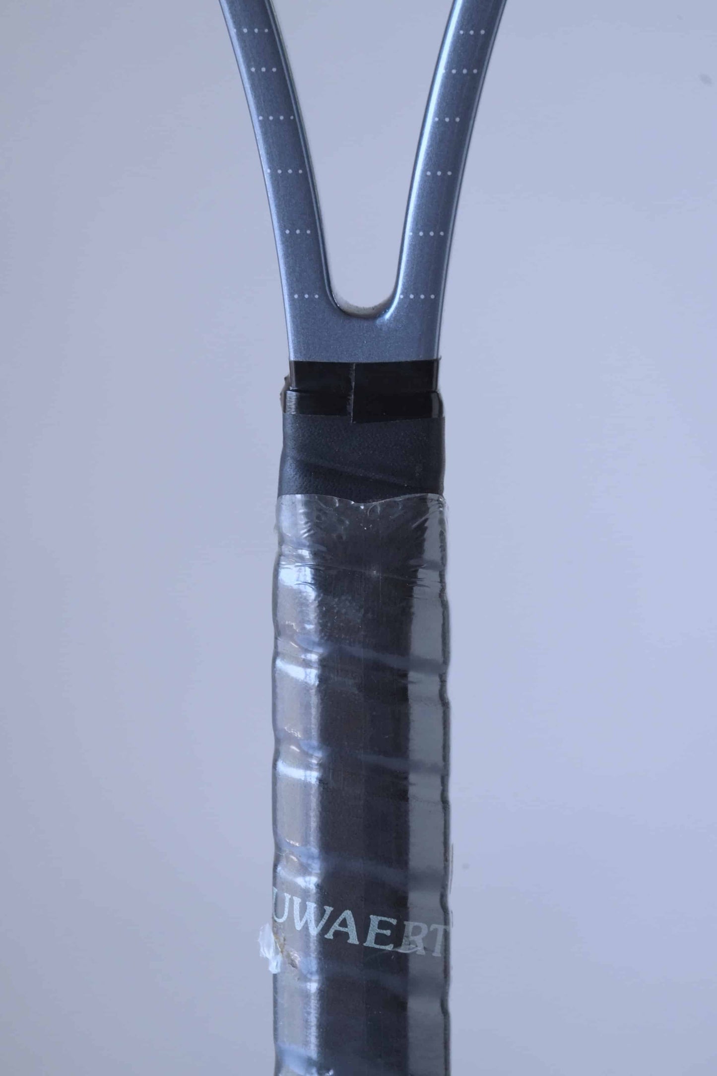 Close up of the racquet handle of a vintage Snauwaert Hi-Ten 30 tennis racket, showing part of the word 'SNAUWAERT' in grey. The handle is wrapped with black overgrip tape and covered with plastic.