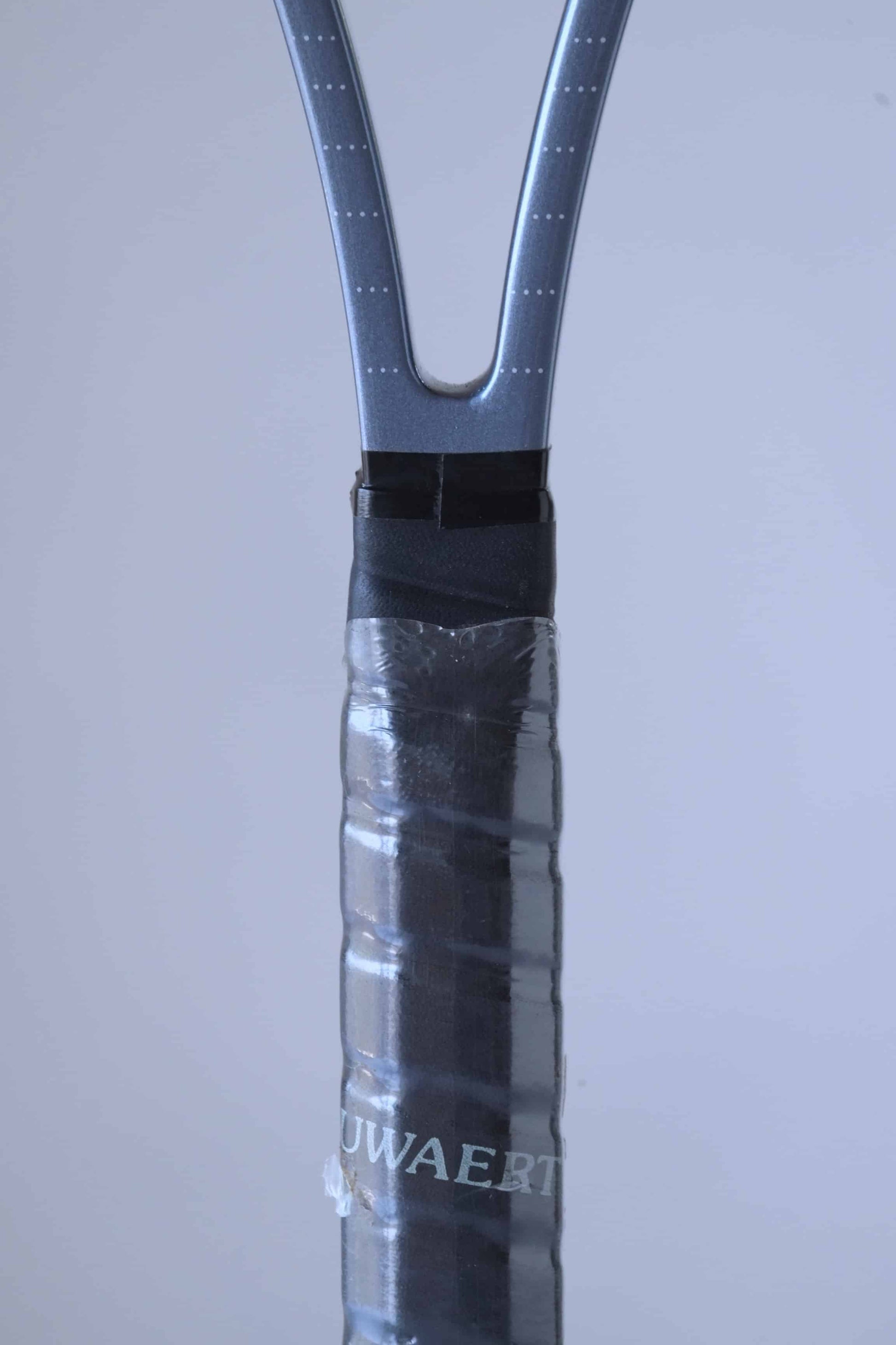 Close up of the racquet handle of a vintage Snauwaert Hi-Ten 30 tennis racket, showing part of the word 'SNAUWAERT' in grey. The handle is wrapped with black overgrip tape and covered with plastic.