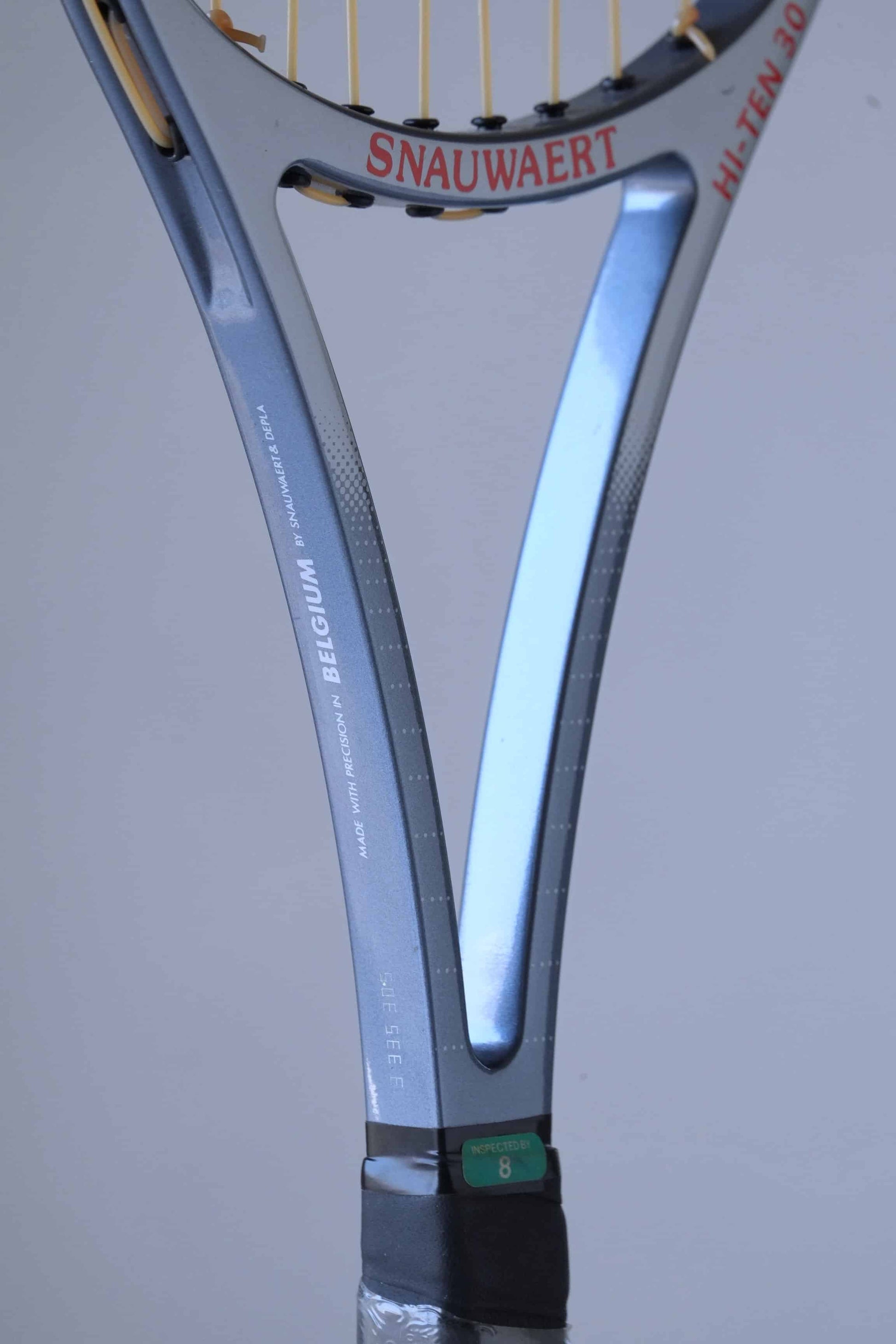 Close-up of a Snauwaert tennis racket handle.
 The handle is blue and black with a textured grip. The word "SNAUWAERT" is written in white on the handle, and the number "8" is visible below it. The racket is made in Belgium.
