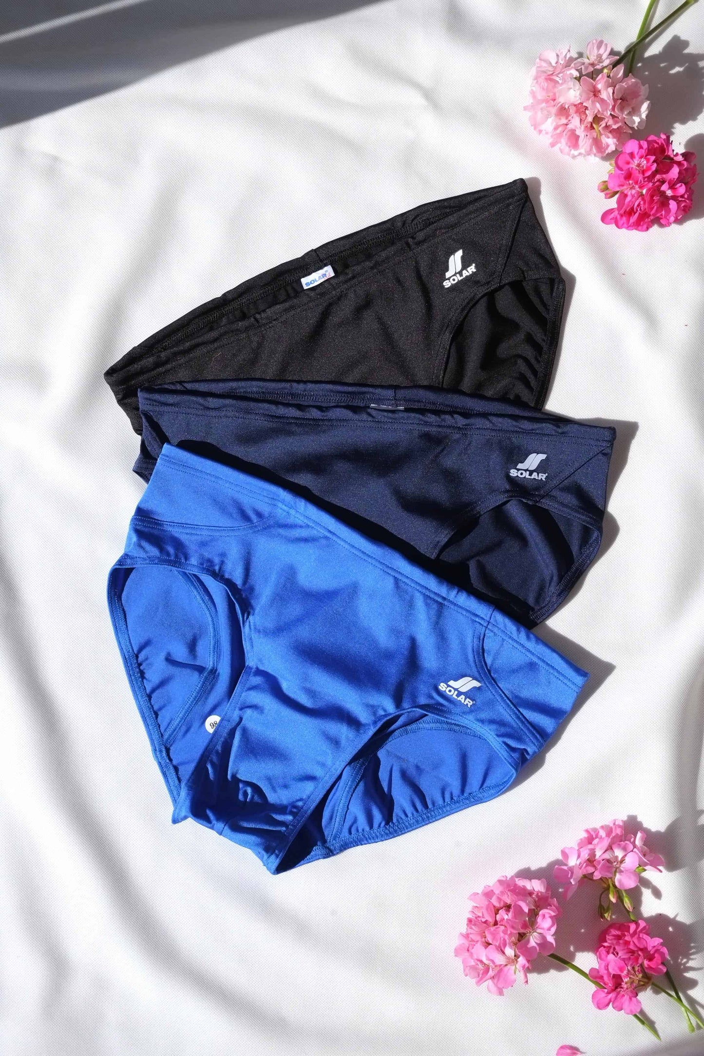 Three SOLAR vintage 70s swim briefs in black, navy, and bright royal blue, laid out under natural light.