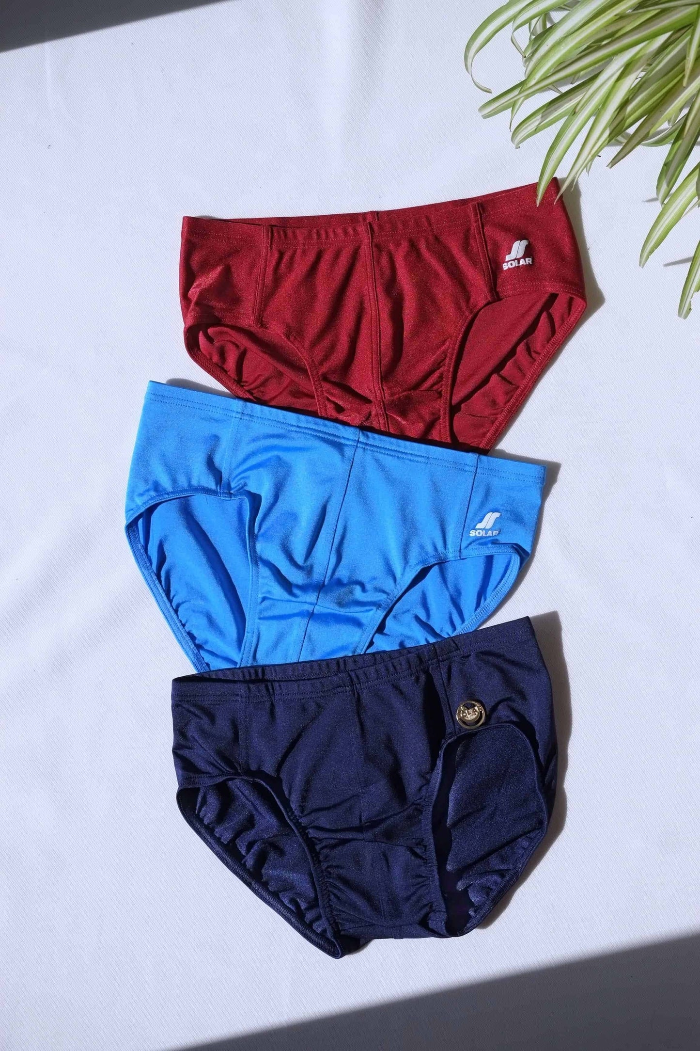 Three SOLAR vintage 70s swim briefs in burgundy red, blue, and navy laid flat under bright lighting.