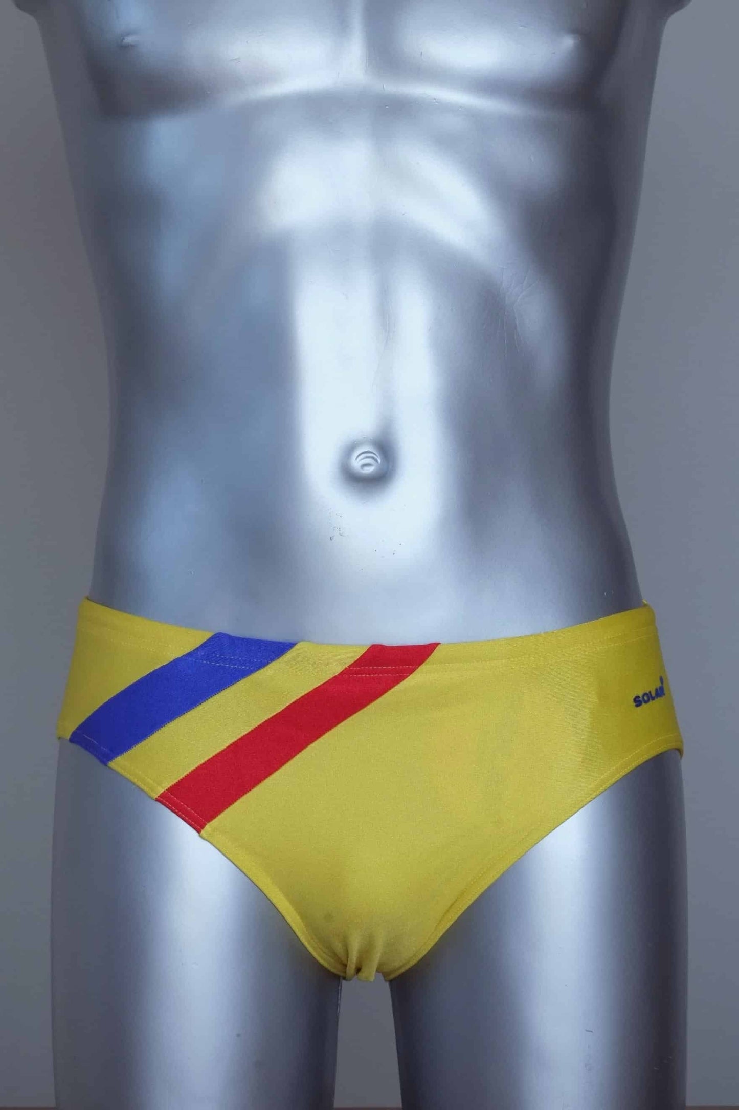 Vintage solar yellow men's swim briefs with red and blue stripes on a silver mannequin against a grey background.