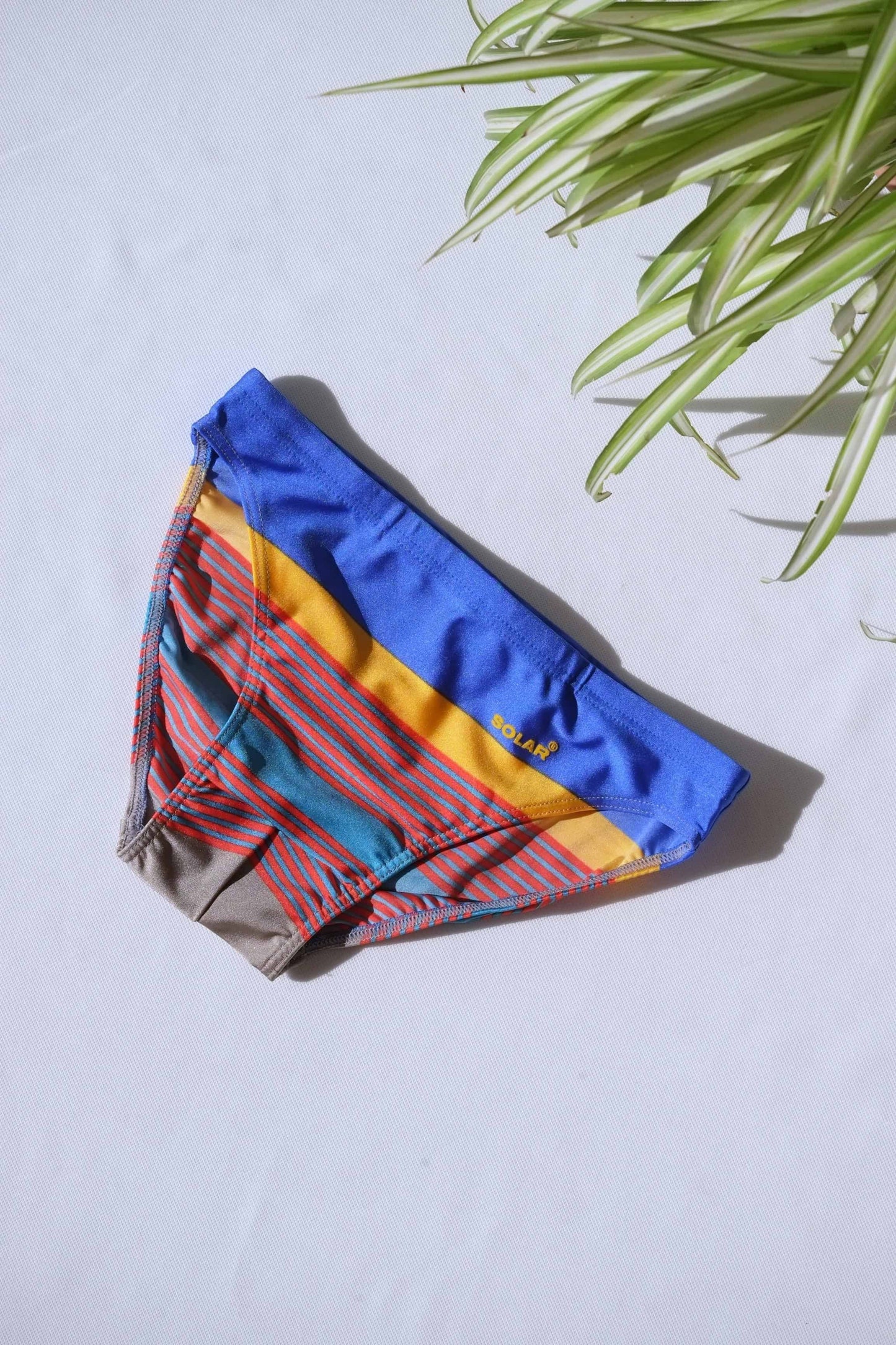 A pair of SOLAR brand vintage 80s men's swim briefs with multi-colored horizontal stripes in blue, yellow, and red hues, laid flat on a white background.
