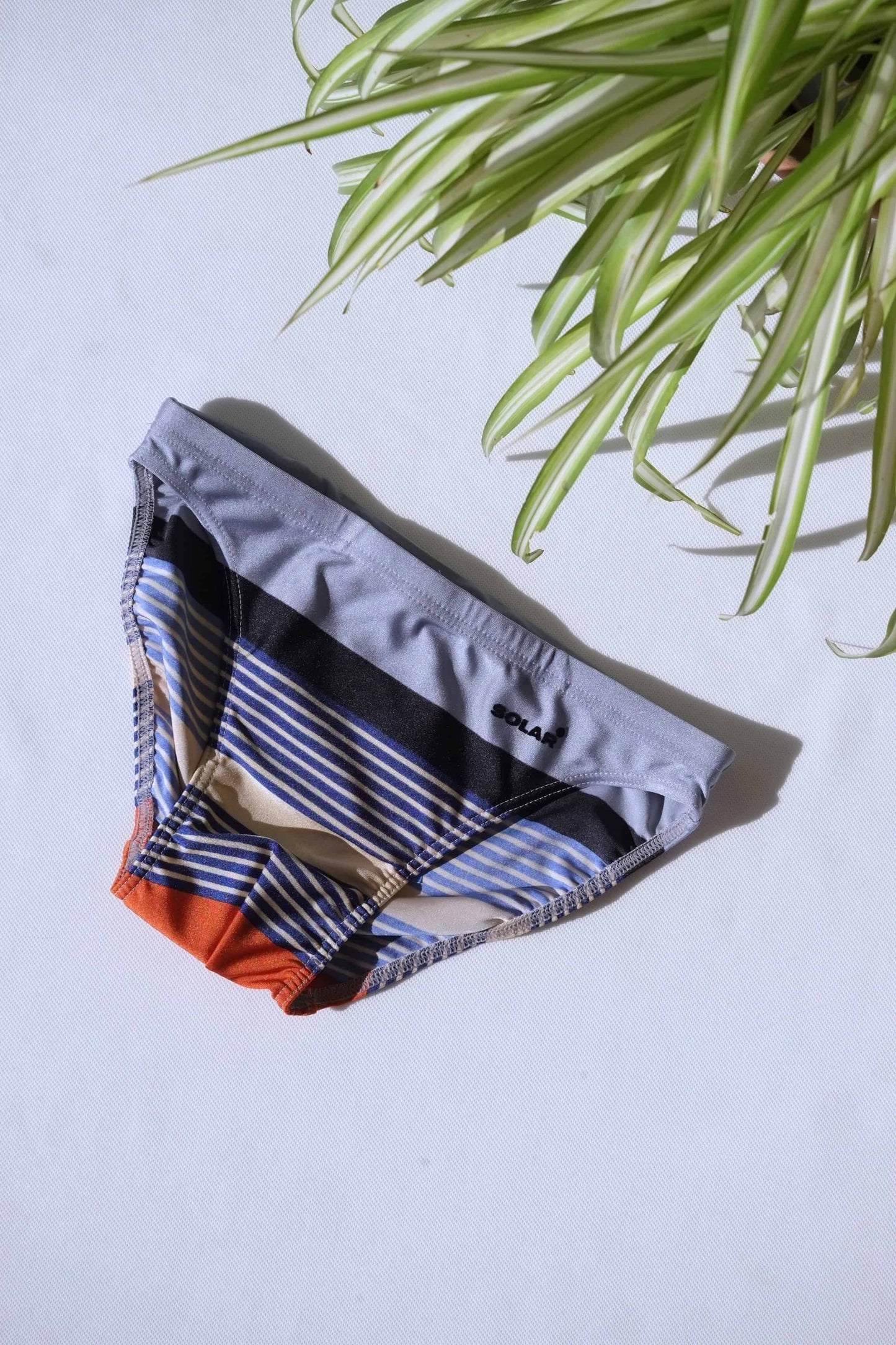 A pair of SOLAR brand vintage 80s men's swim briefs with multi-colored horizontal stripes in grey, black, and orange tones, laid flat on a white background.