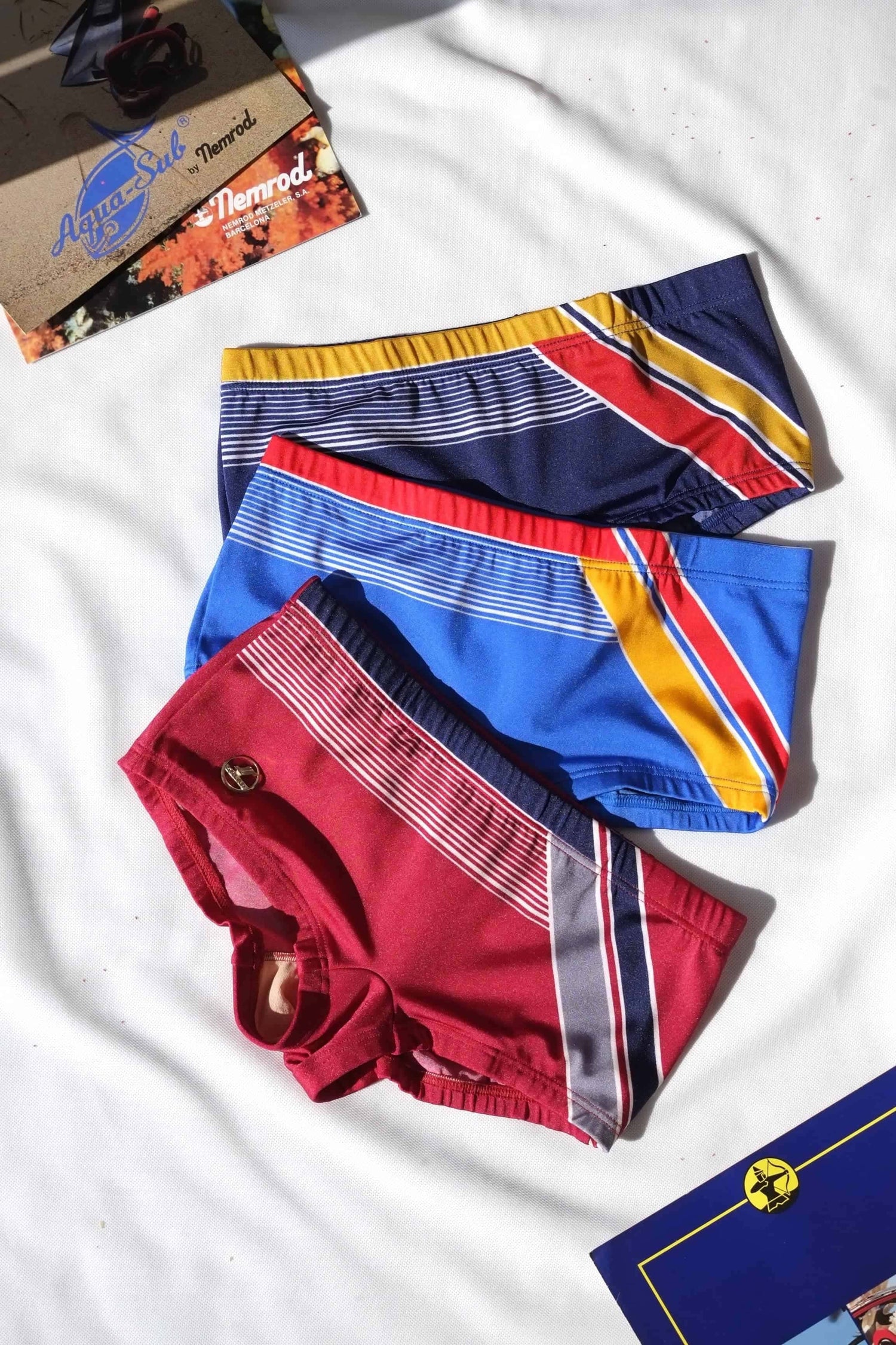 Three pairs of colorful swim shorts on a white background with magazines in the background.